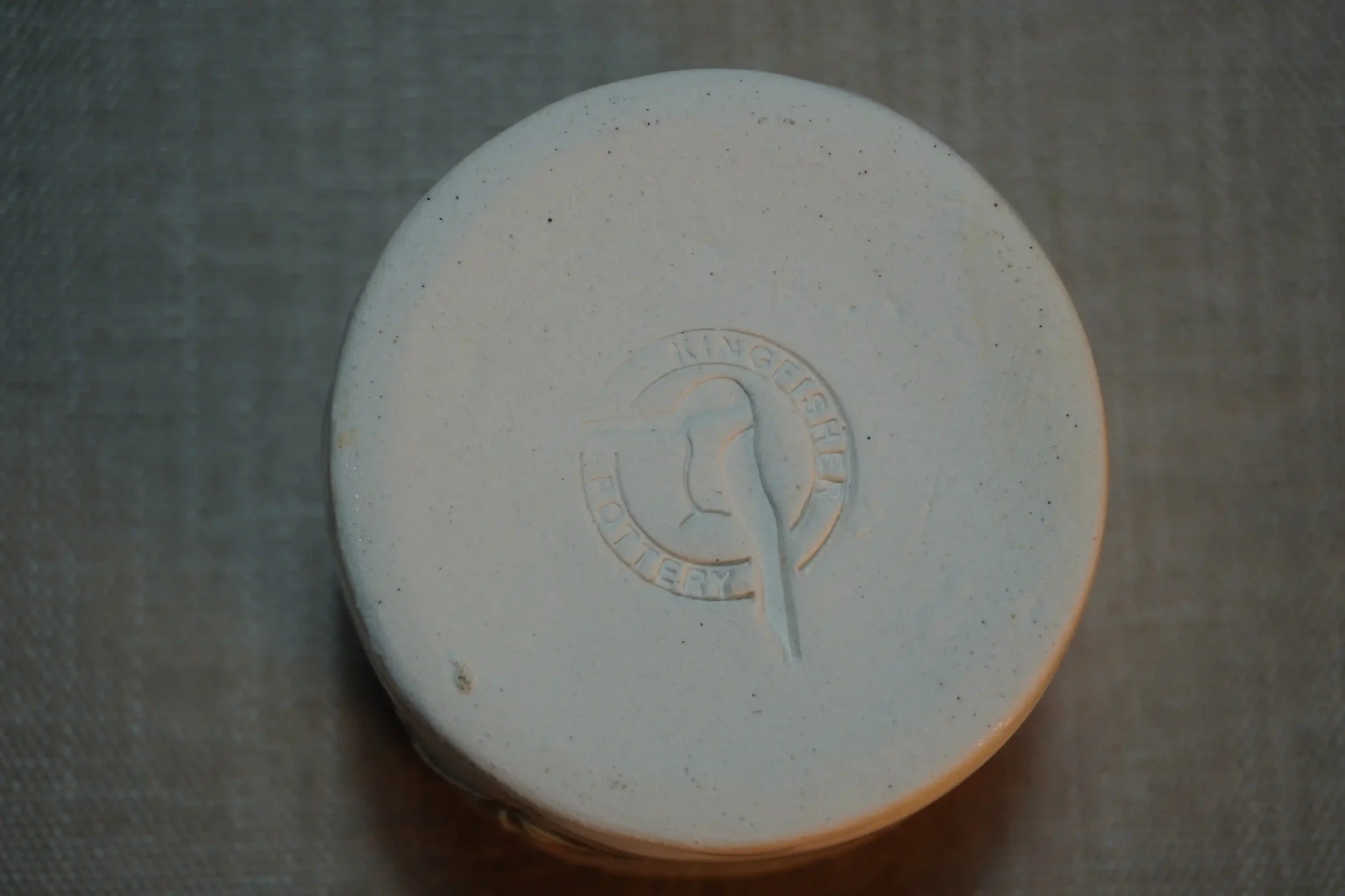 View of a pottery maker's mark on the bottom of a ceramic pincushion. The maker's mark shows a kingfisher and words saying Kingfisher Pottery.