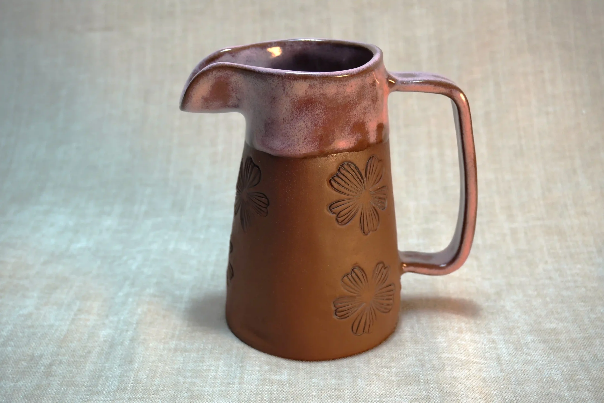 A terracotta brown ceramic pitcher with pink glazed upper and floral design, set against a neutral background.