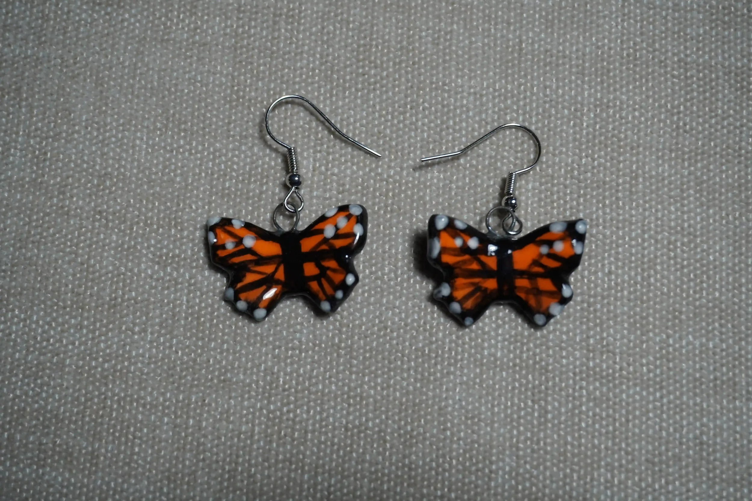 Pair of Ceramic monarch butterfly-shaped earrings with orange, black, and white pattern, placed on beige textured surface.