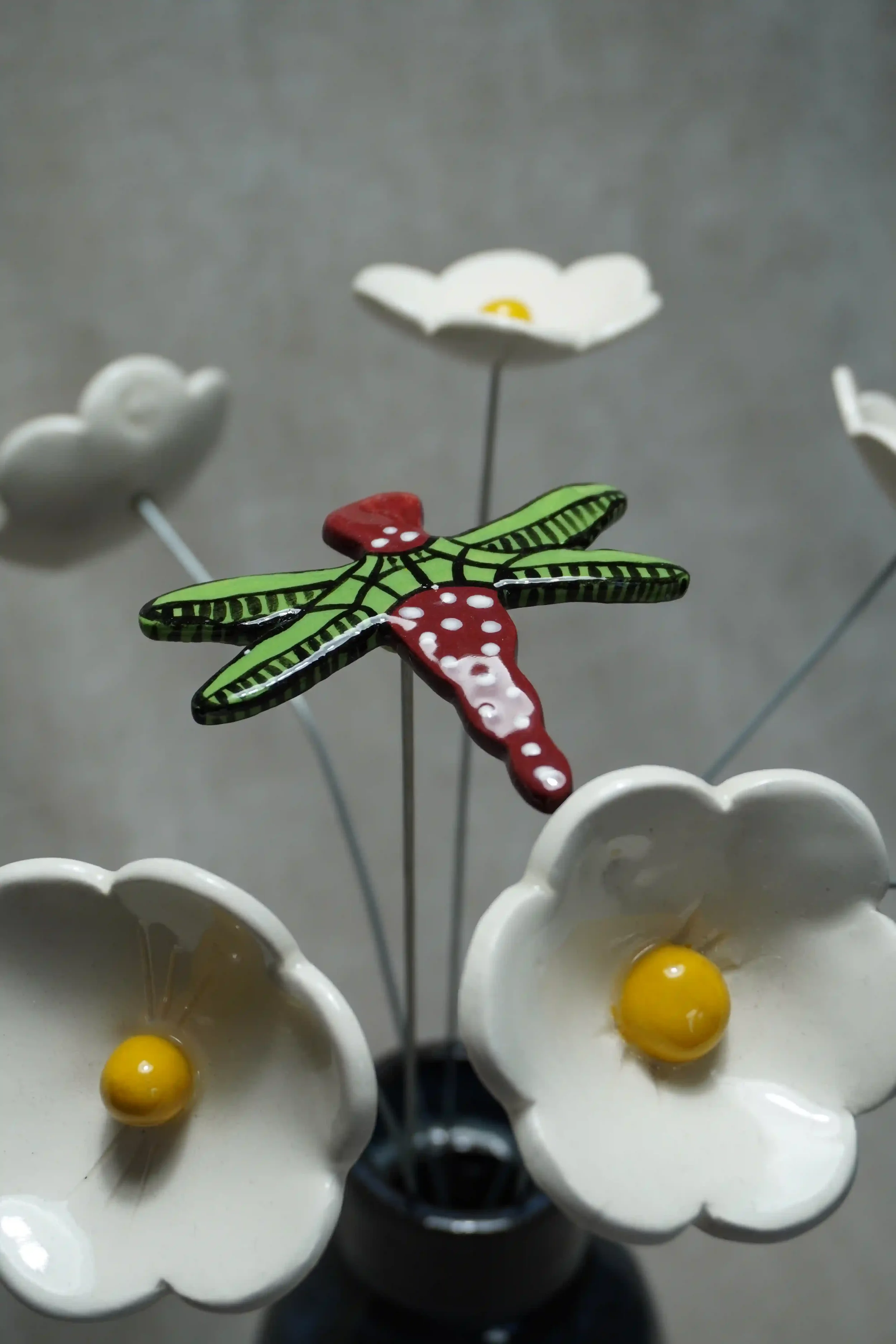 A close-up of a colorful ceramic dragonfly with green and black wings and a red body with white speckles, set amongst white ceramic daisies with yellow centres. All mounted on thin metal stems, against a plain gray background
