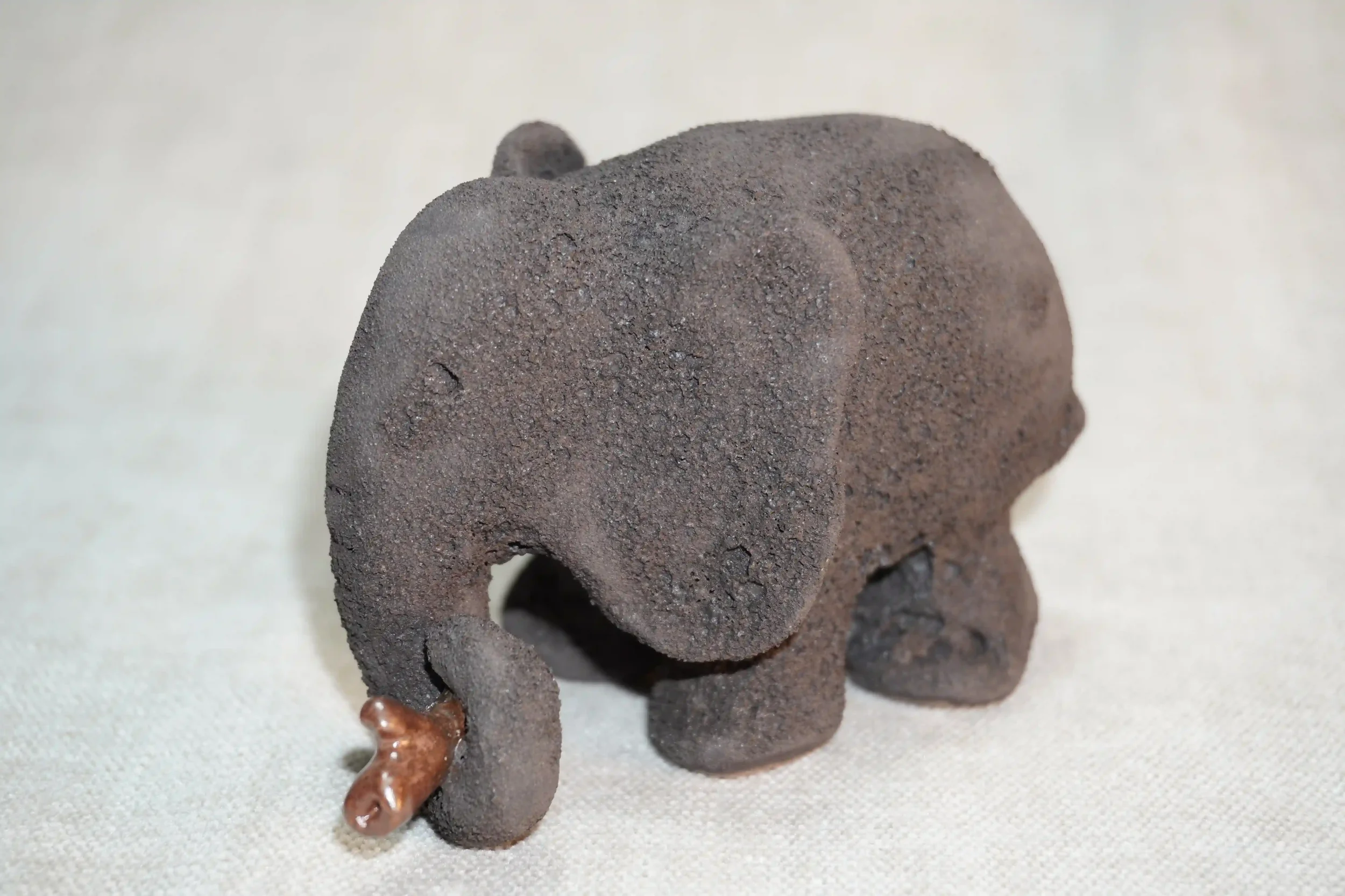 Close-up view of a handmade pottery elephant figurine with a dark grey textured surface and holding a branch in it's trunk, placed on a light-colored fabric surface.