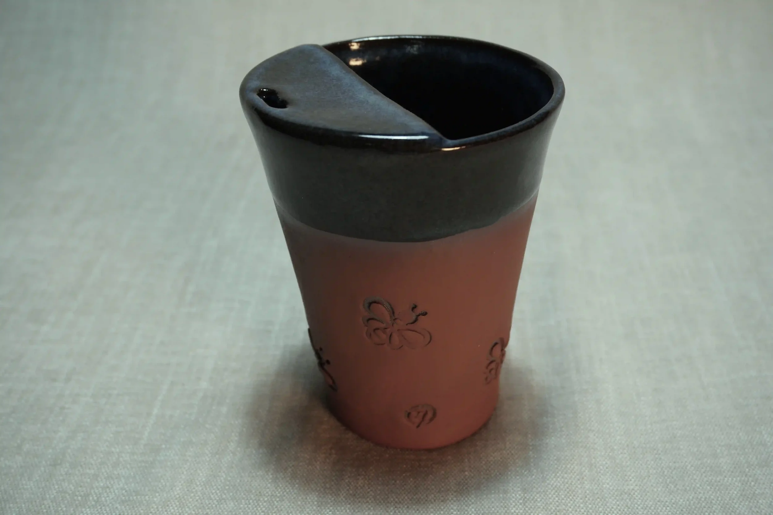 A ceramic travel mug with a dark blue glaze on the inside, a terracotta brown base, and a decorative floral pattern on the outside, placed on a light-colored surface.