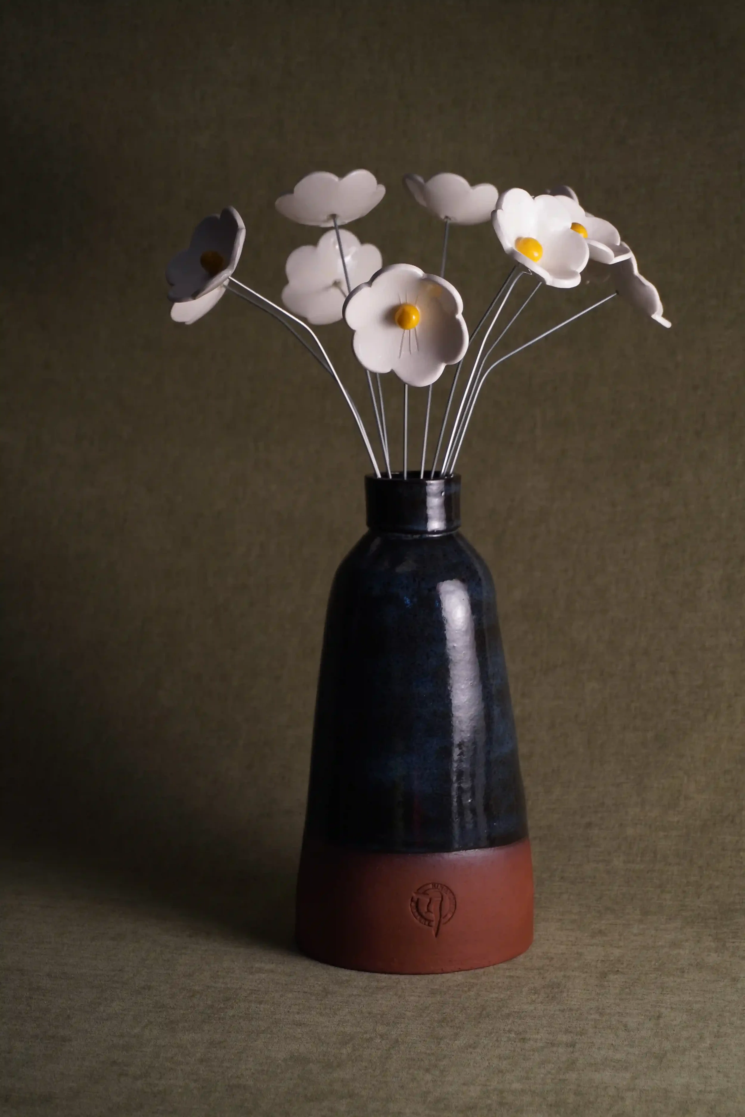 A bouquet of white ceramic Daisy flowers with yellow centers in a blue glazed and terracotta-colored ceramic vase.
