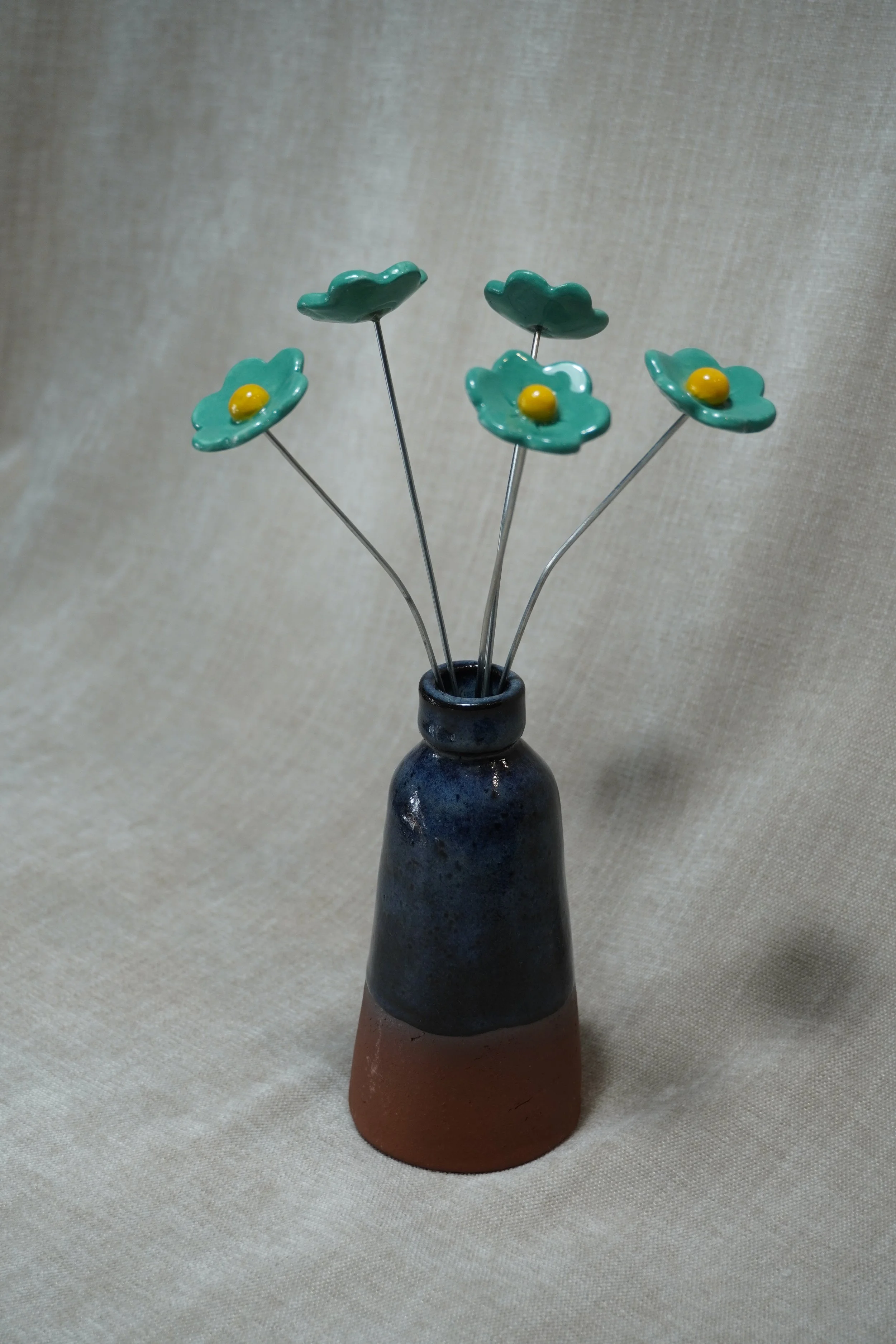A ceramic pottery vase with a wide, rounded terracotta base, narrow neck, and a blue glaze, holding five ceramic green flowers with metal stems, displayed on a light neutral background.