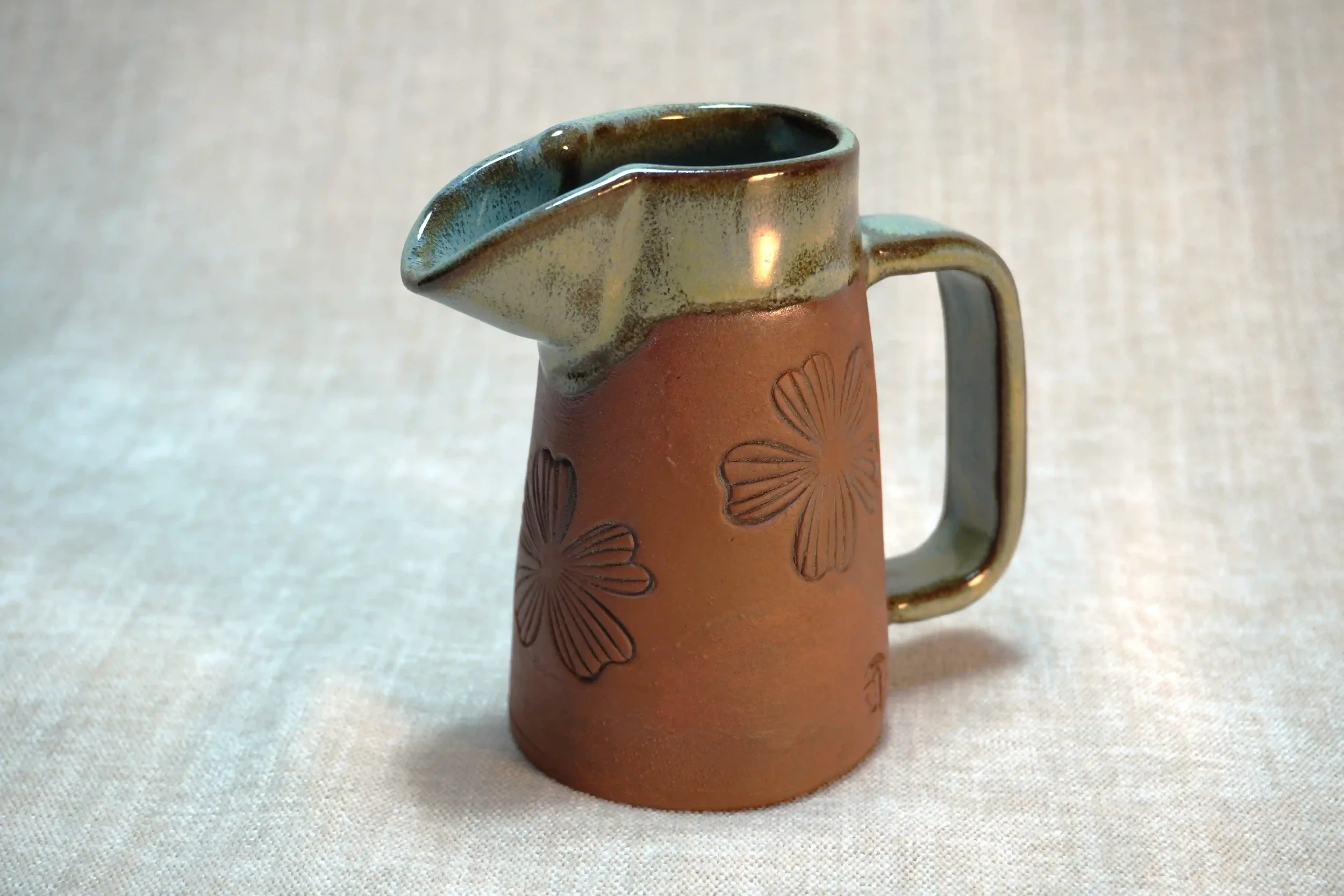 A small ceramic pitcher with embossed floral design on the terracotta-colored body, glazed in light green, with a handle on the side and a spout on top. 
