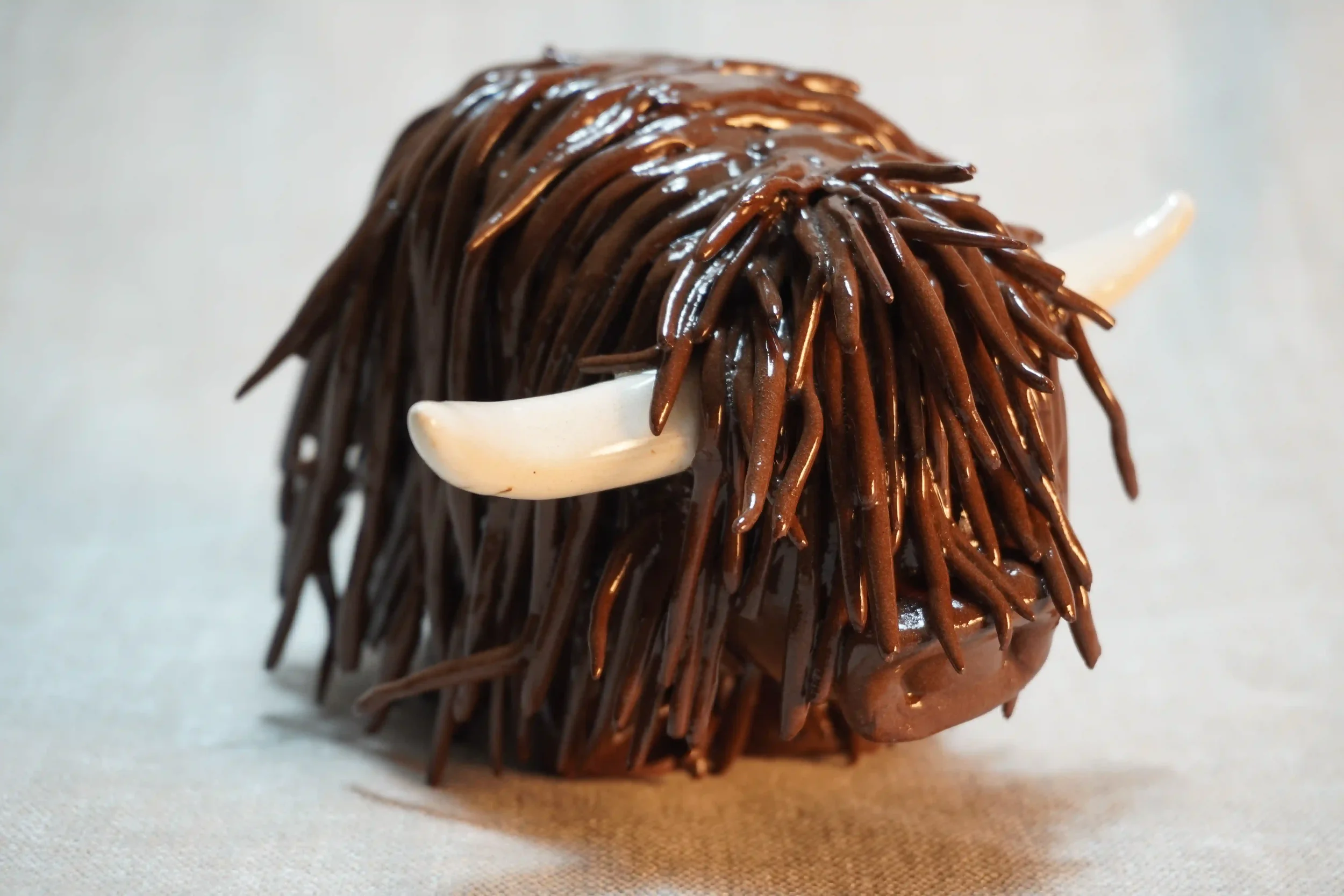 Quarter front view of a handmade pottery ceramic sculpture of a Highland Cow ornament with deep brown-coloured hair and beige horns.