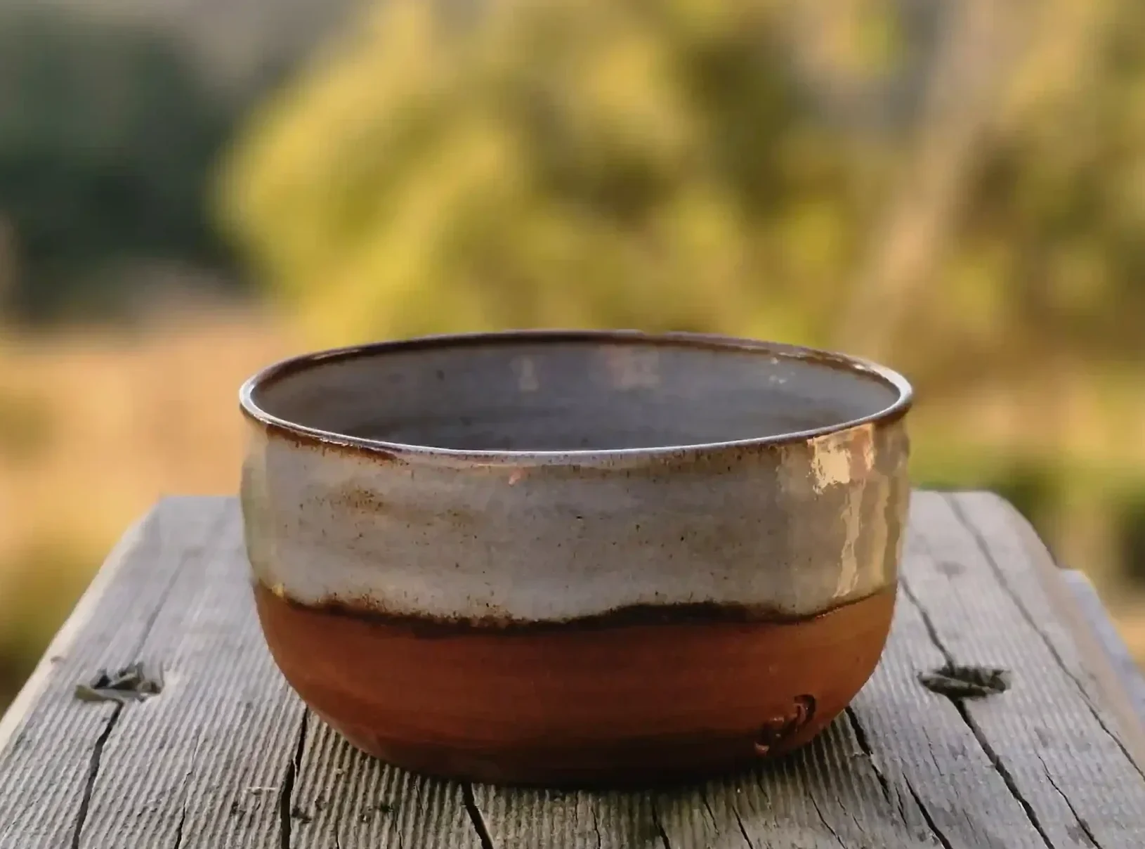 A ceramic bowl with a creamy grey-white glaze on the top and a terracotta unglazed bottom, sitting on a weathered wooden surface outdoors with a blurred background of green foliage.