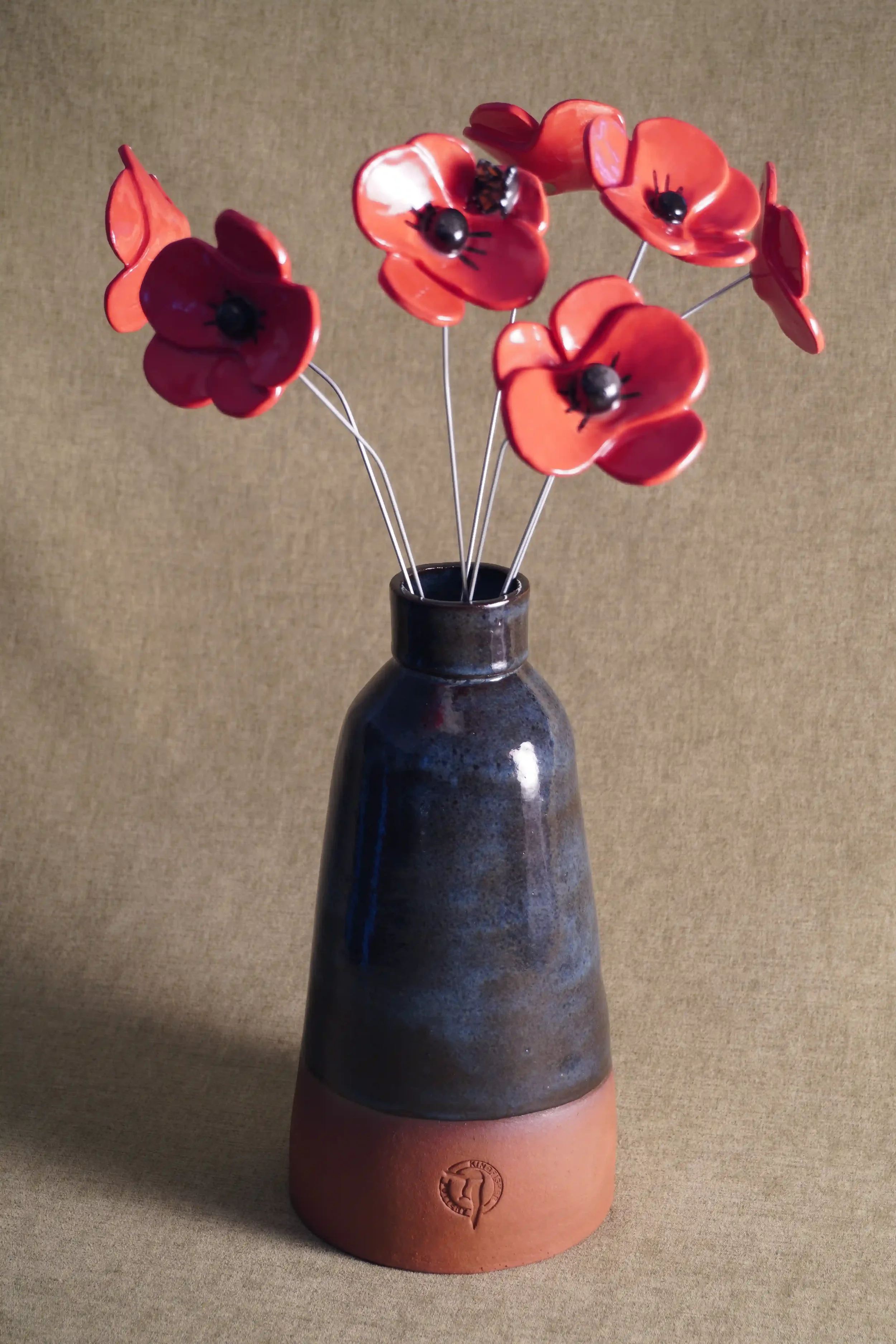 A blue-glazed hand-made ceramic vase with terracotta base, holding seven spectacular red ceramic poppy flowers with black centers on thin wire stems, set against a textured brown background.