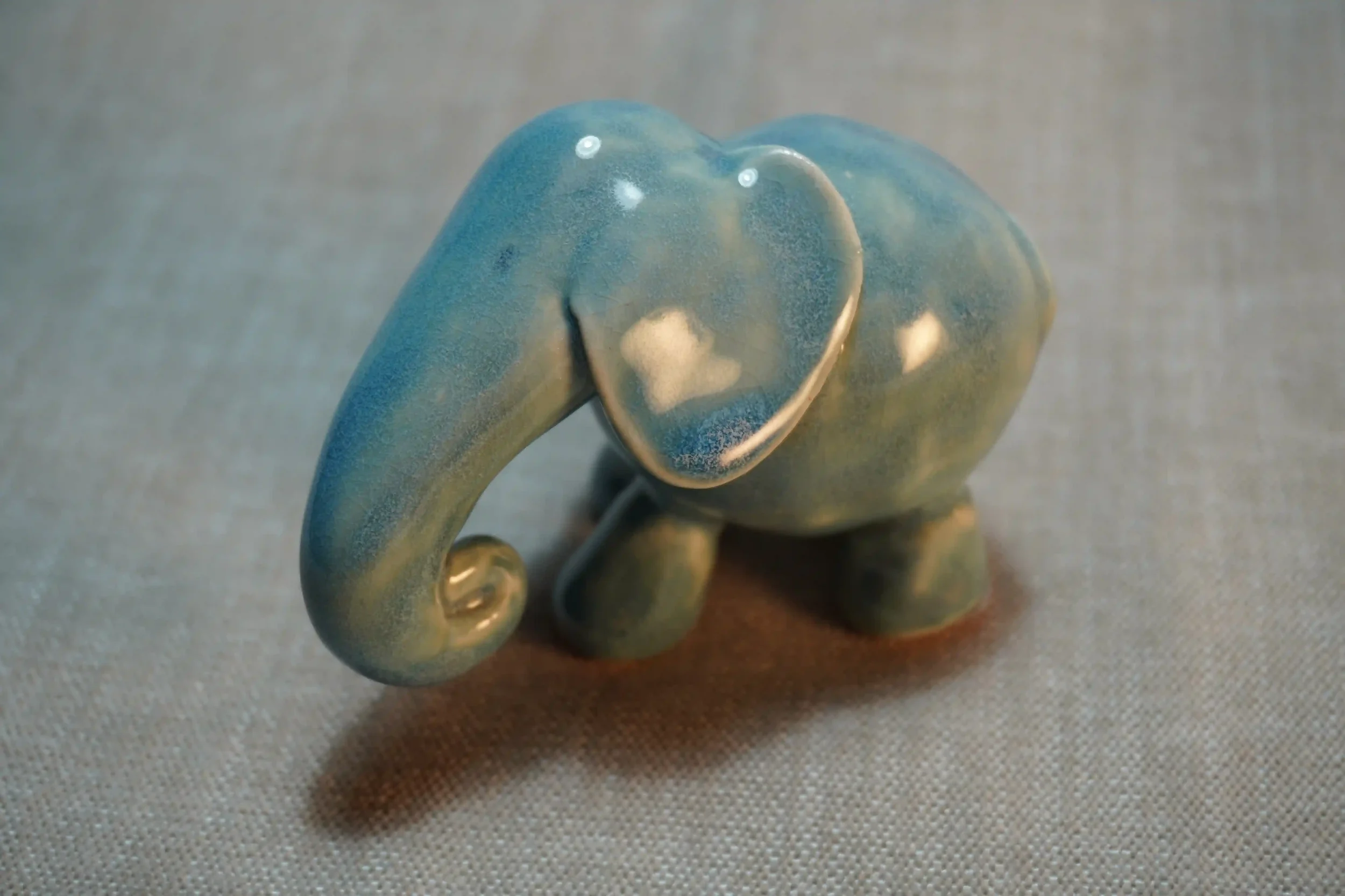 A small, decorative elephant sculpture made of glazed ceramic with a blue-green finish, sitting on a textured surface.