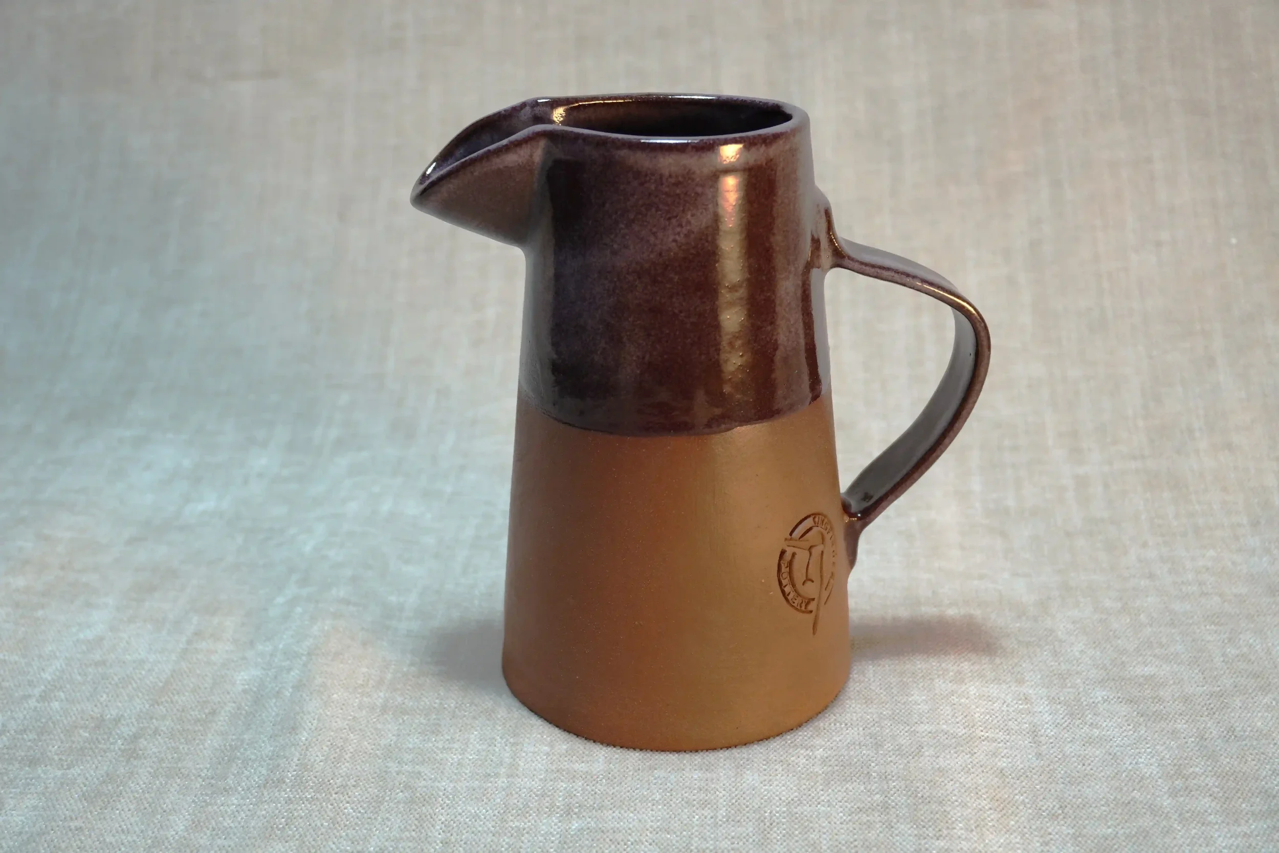 A ceramic pitcher with terracotta brown colour and purple glaze and the Kingfisher Pottery logo embossed near the bottom, standing on a plain, light-colored fabric surface.