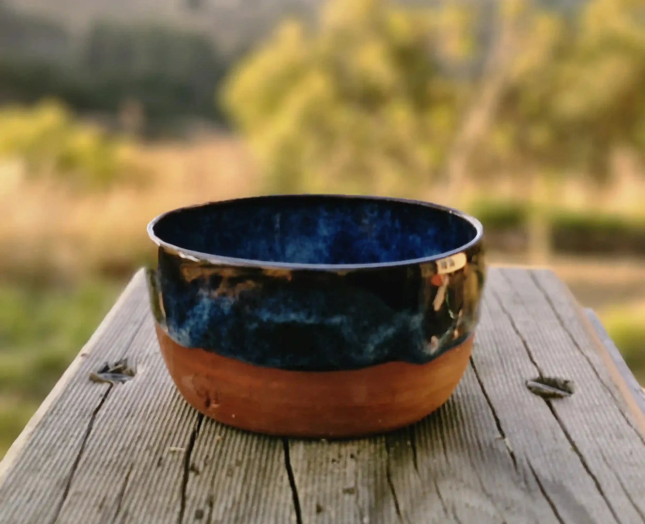 Close-up of a ceramic pottery bowl with a blue glazed upper and terracotta brown base on a wooden outdoor surface, with a blurred natural background.