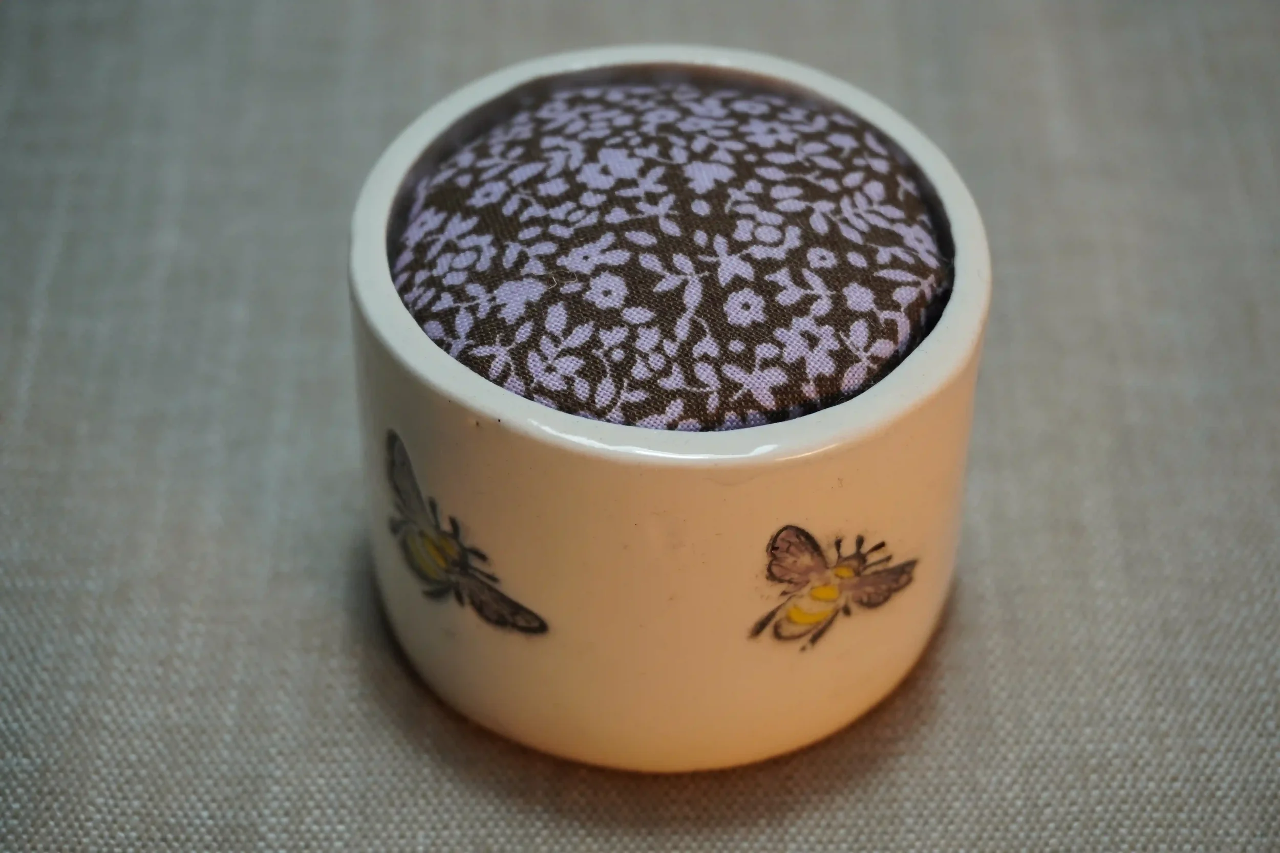 A ceramic pincushion with bee design, filled with a brown and pink floral patterned cushion. The pincushion is shown on a textured surface.