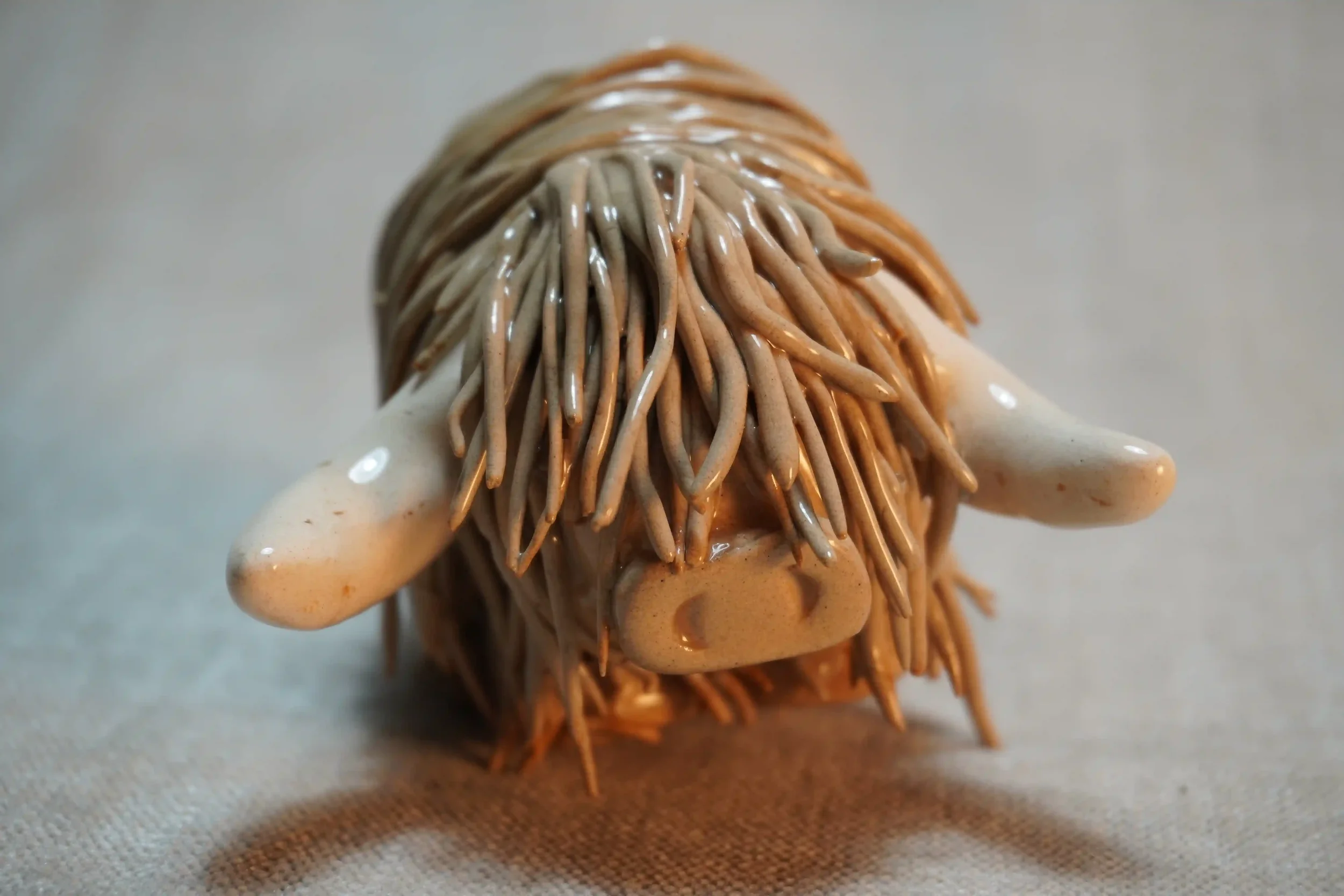 Frontal view of a handmade pottery ceramic sculpture of a Highland Cow ornament with light brown-coloured hair and beige horns.