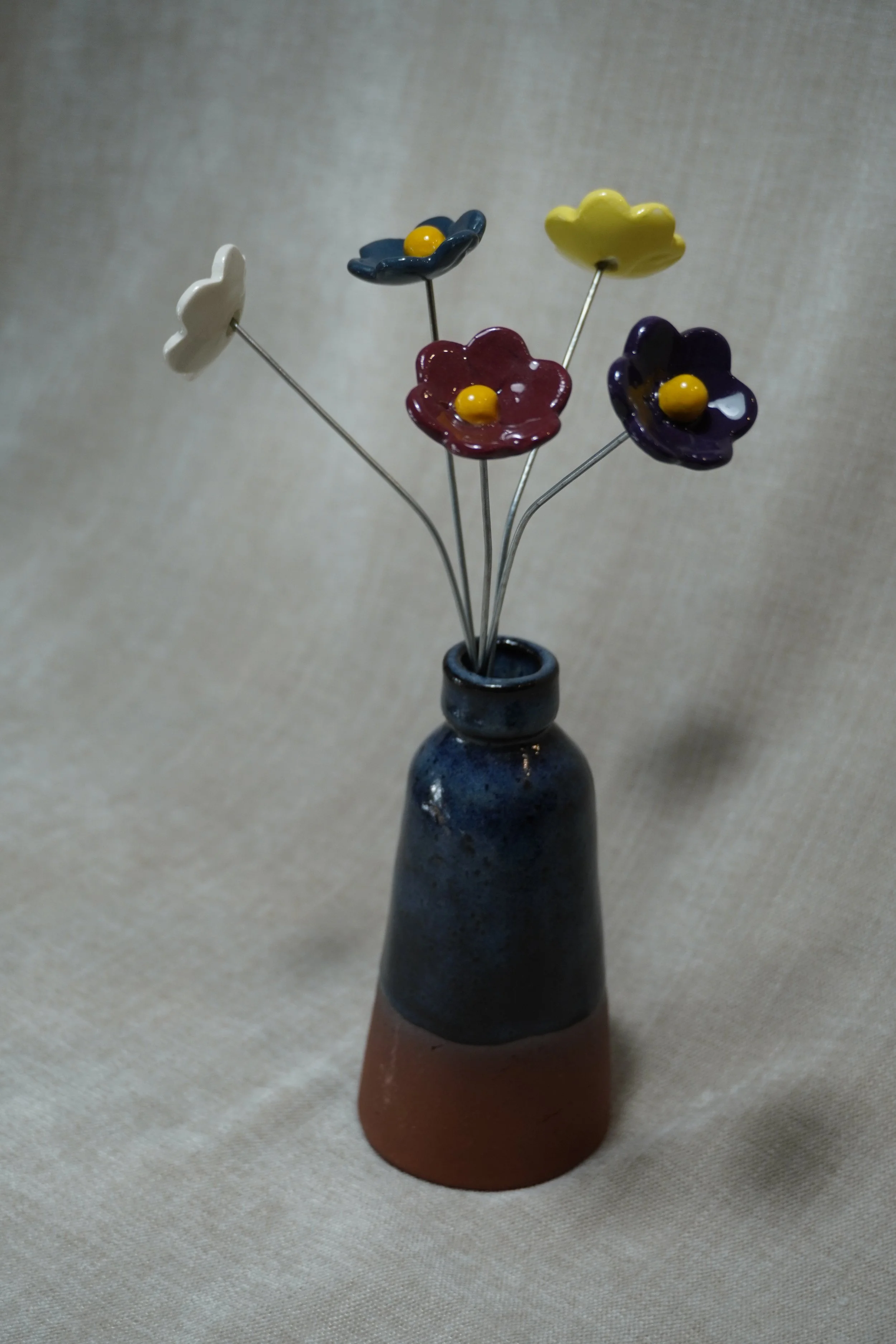 A ceramic vase with a wide, rounded terracotta base, narrow neck, and a blue glaze, holding five ceramic flowers, in white, maroon, purple, blue and yellow, with yellow centres and metal stems, displayed on a light beige background.