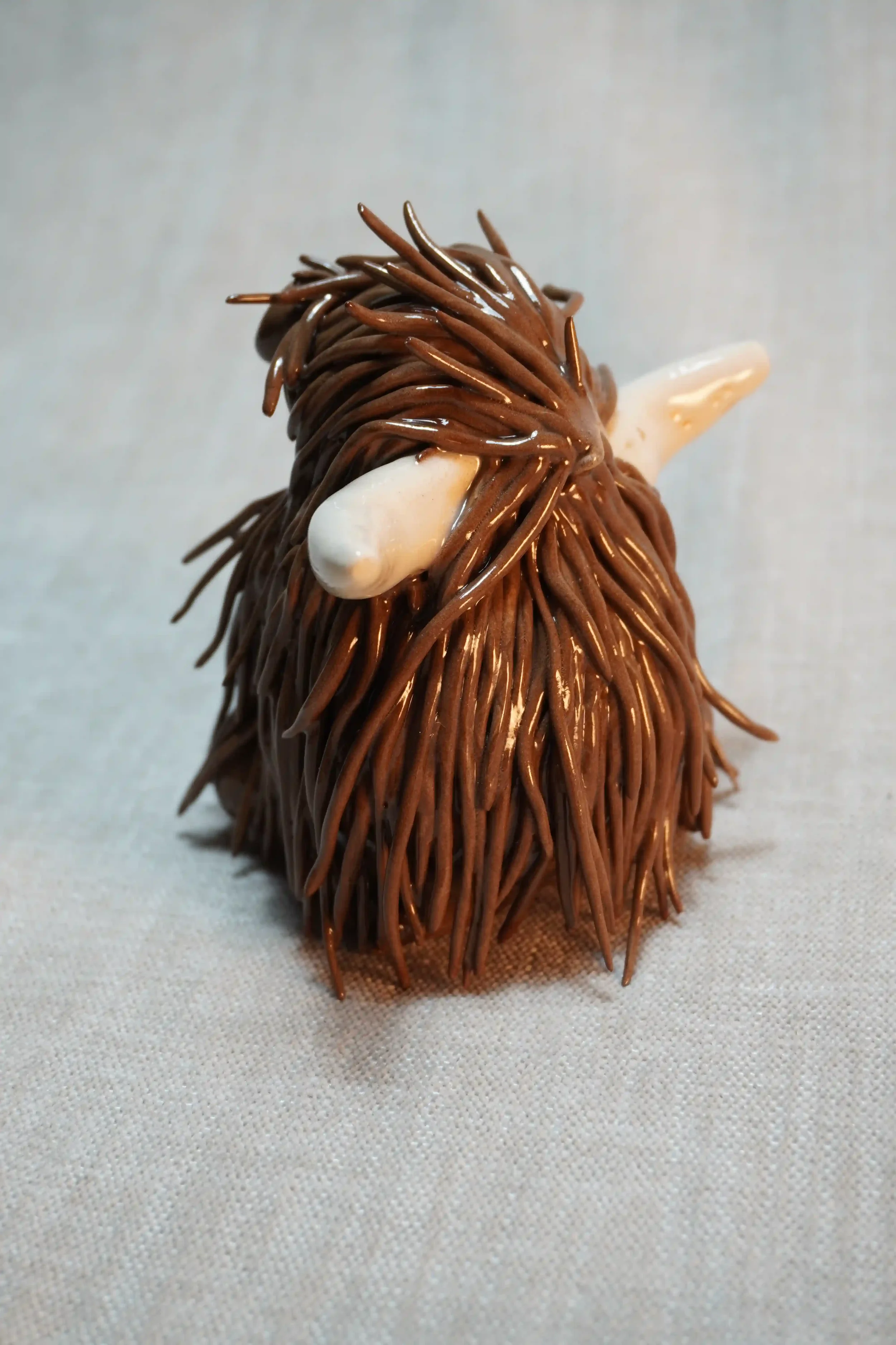 Quarter rear view of a handmade ceramic pottery sculpture of a Highland Cow ornament with rich dark chocolate brown-coloured hair and beige horns.