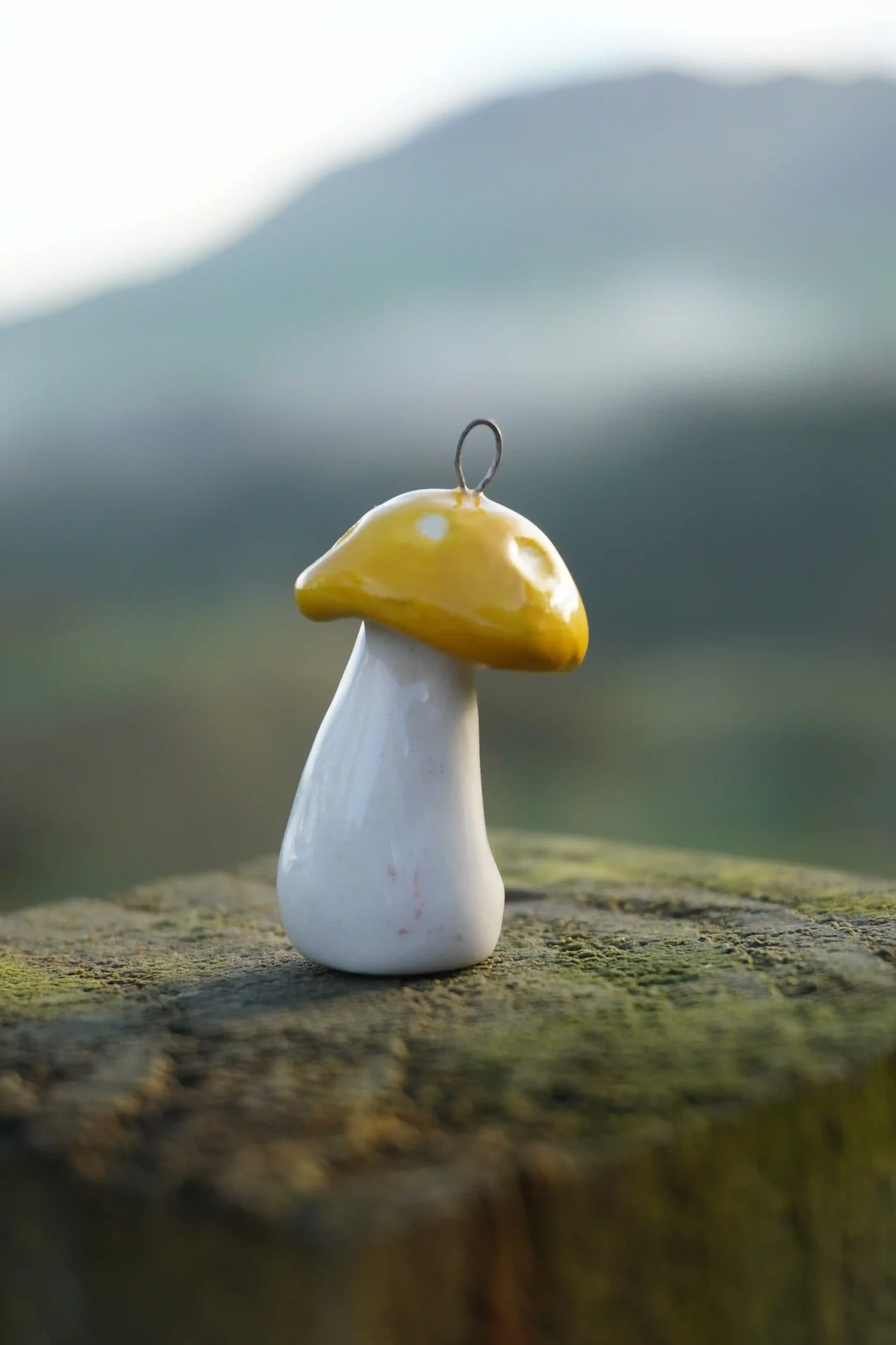 Close-up of a ceramic mushroom ornament with a yellow cap and white stem, placed on a weathered wooden surface outdoors, with a blurred background of sky and landscape.