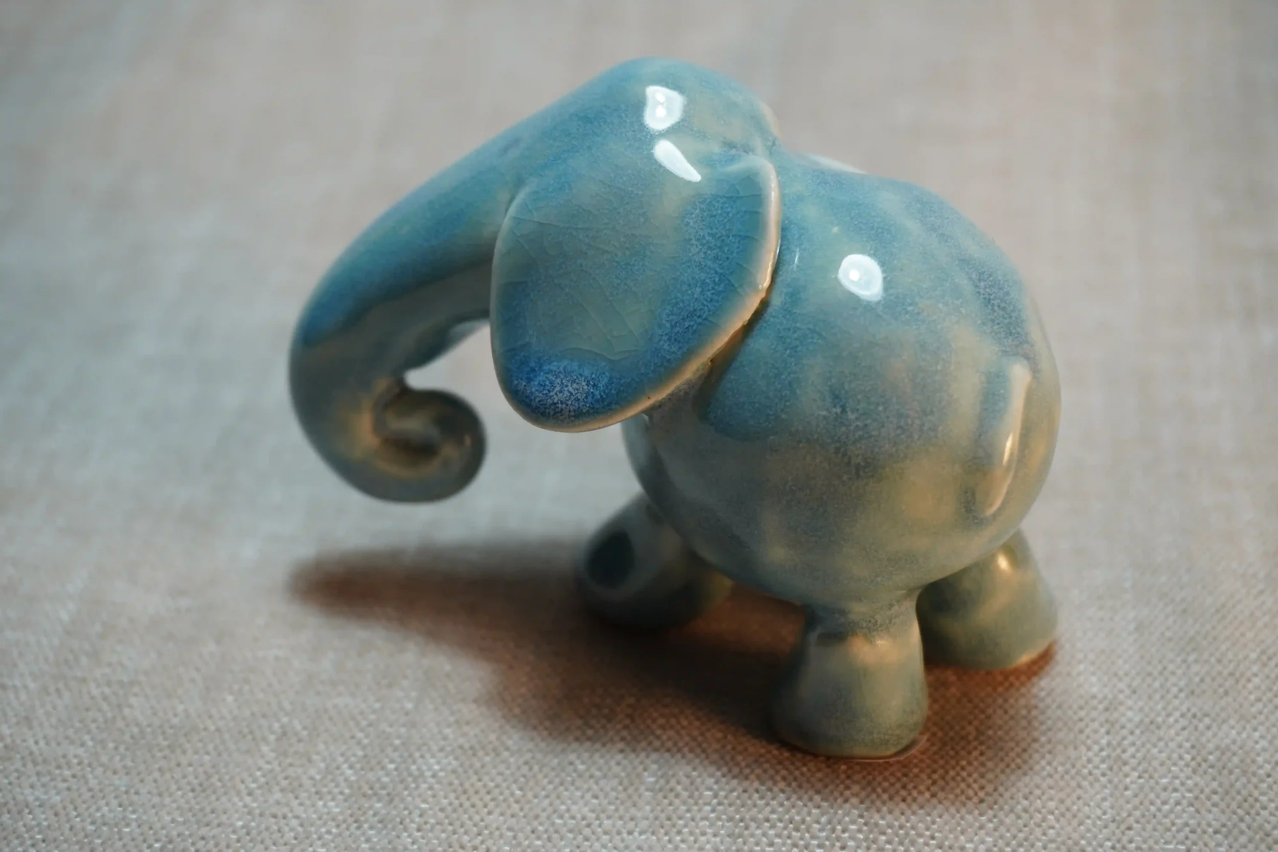 A small, handmade ceramic figurine of an elephant with a blue glazed finish, positioned on a textured light-colored surface.