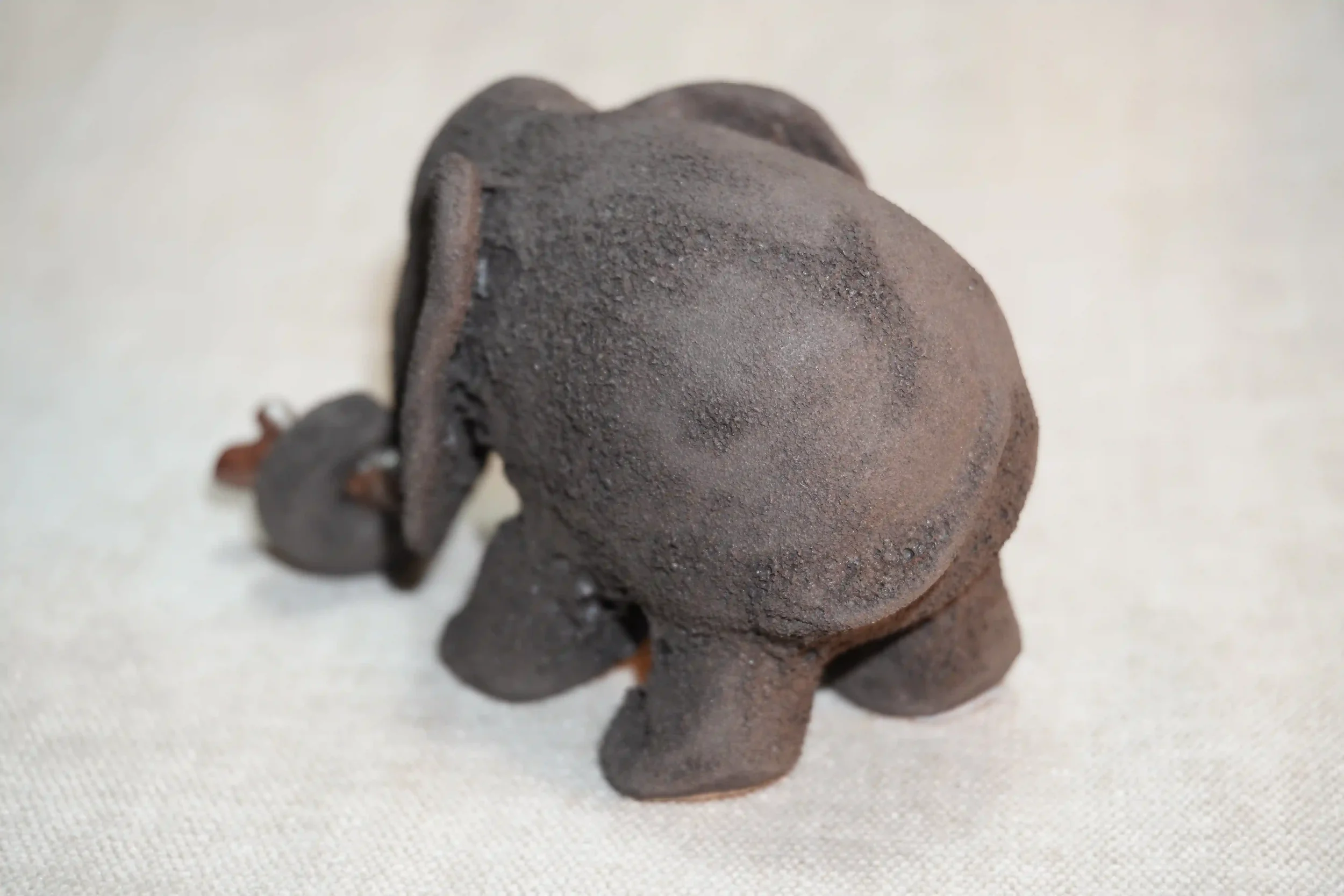 Rear view of a handmade pottery elephant figurine with a dark grey textured surface and holding a branch in it's trunk, placed on a light-colored fabric surface.