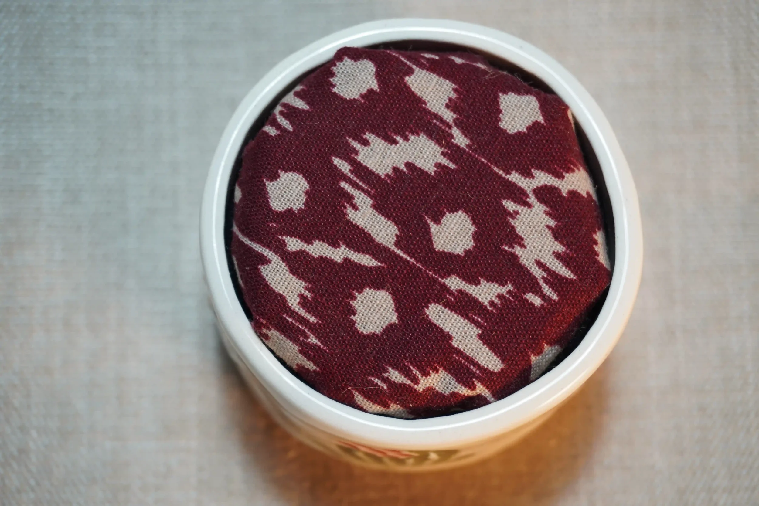 View from above of a ceramic pincushion with a maroon and white patterned fabric-covered cushion. The pincushion is shown on a textured surface.