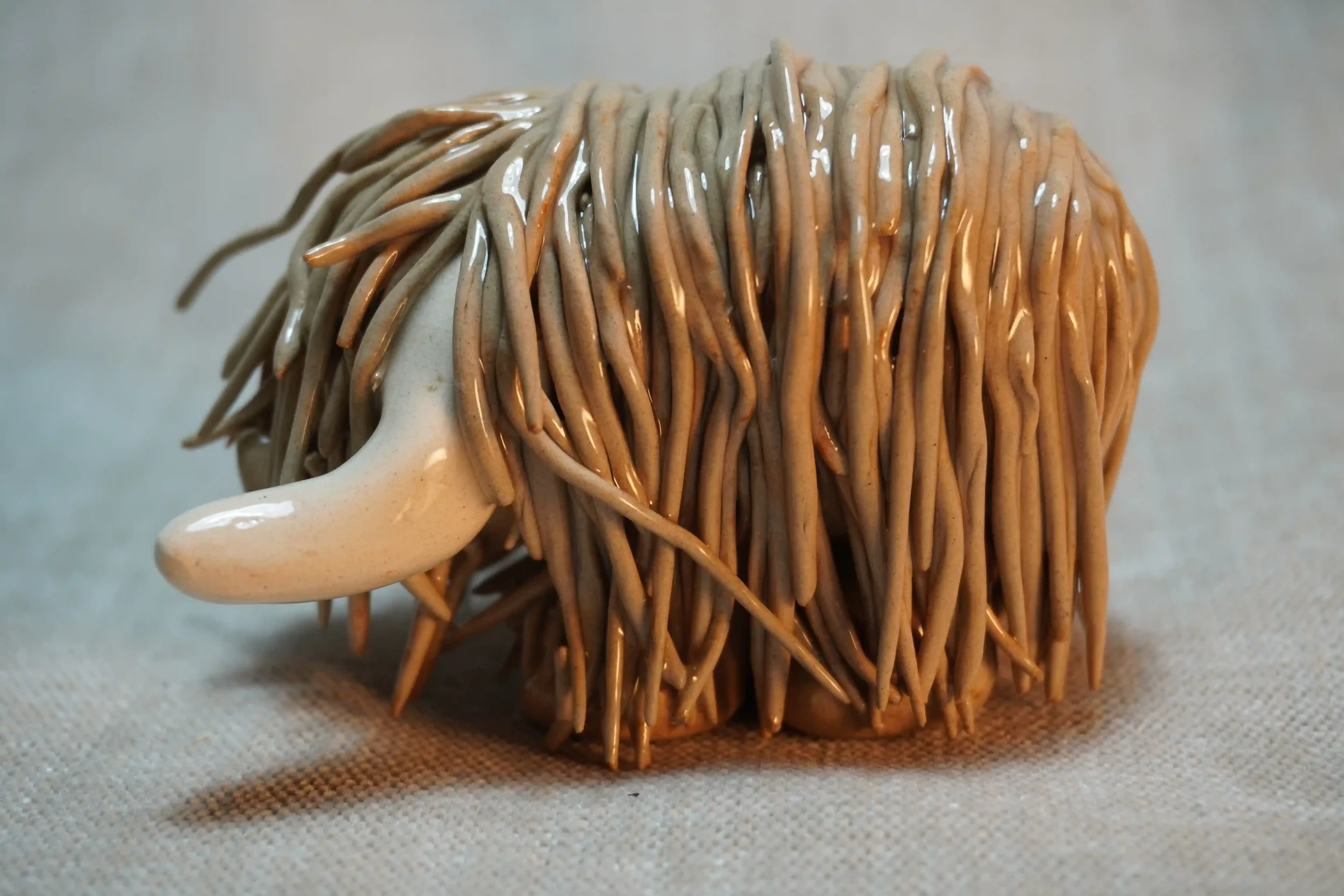 Side view of a handmade pottery ceramic sculpture of a Highland Cow ornament with light brown-coloured hair and beige horns.