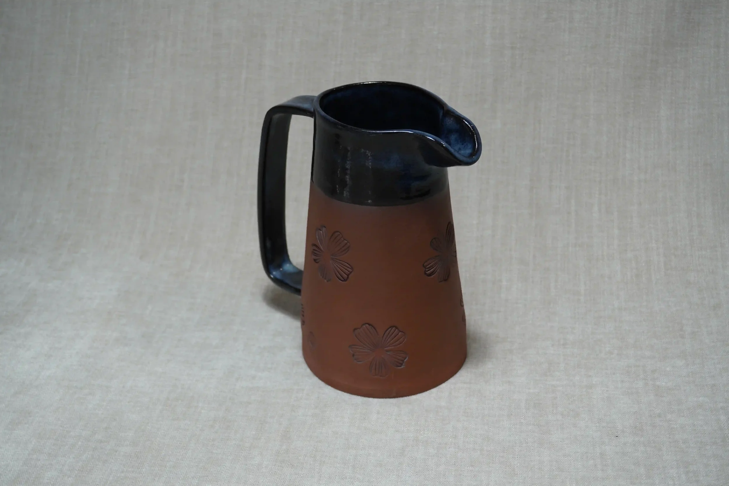 A handmade ceramic pitcher (jug) with a black glazed upper, handle and spout, decorated with floral patterns on its reddish-brown terracotta body, set against a plain background.