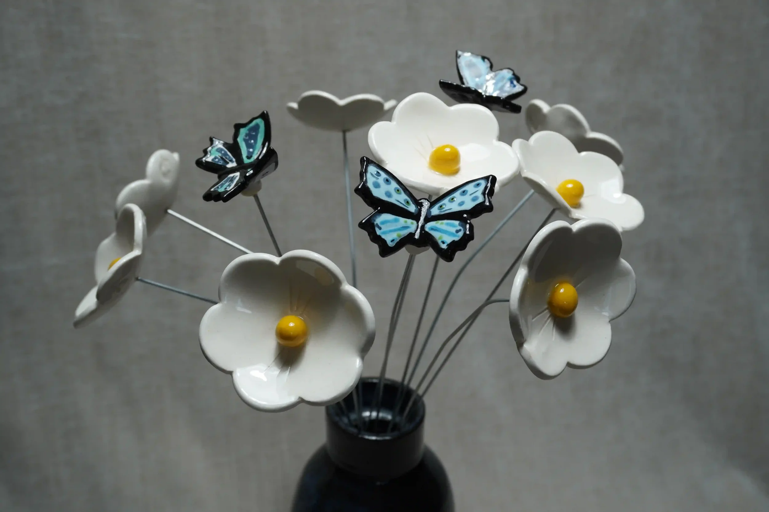 Three blue and black ceramic butterflies on metal stems displayed with white ceramic Daisy flowers with white petals, yellow centers, in a blue-glazed ceramic vase.