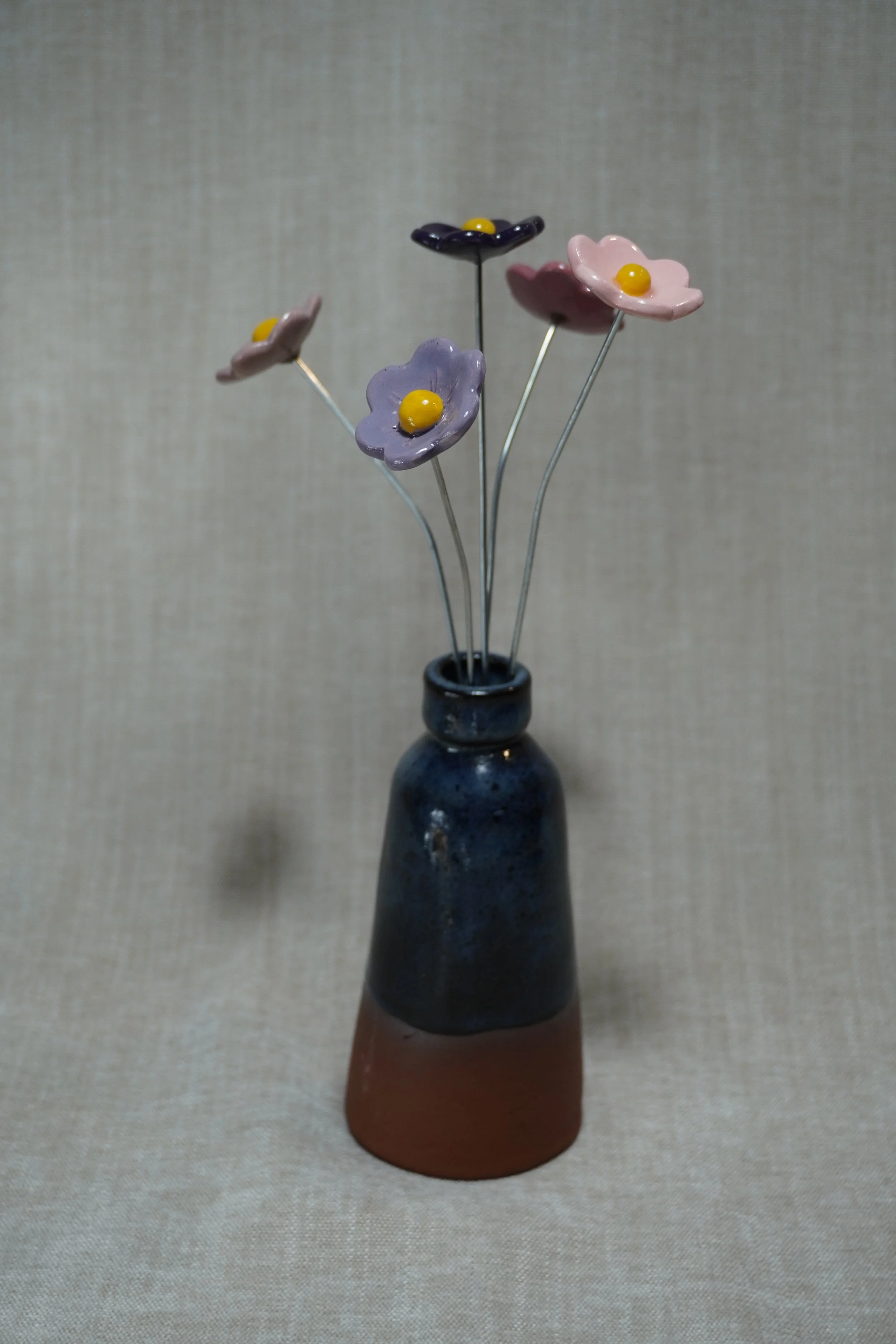 A ceramic pottery vase with a wide, rounded terracotta base, narrow neck, and a blue glaze, holding five ceramic purple, pink and blue flowers with metal stems, displayed on a light neutral background.