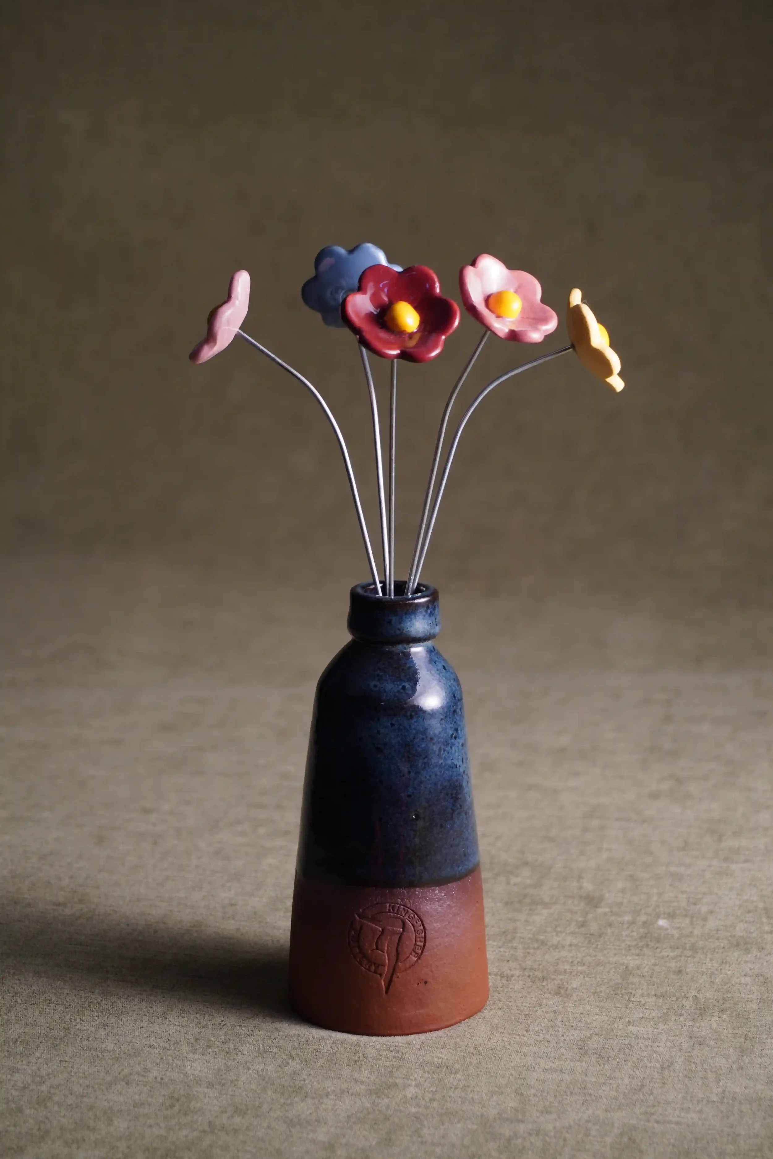 A ceramic vase with a dark glaze on top and a terracotta bottom, holding five wire flower stems with colorful ceramic flowers in pink, red, blue, and yellow. The flowers are arranged in a small bouquet against a neutral background.