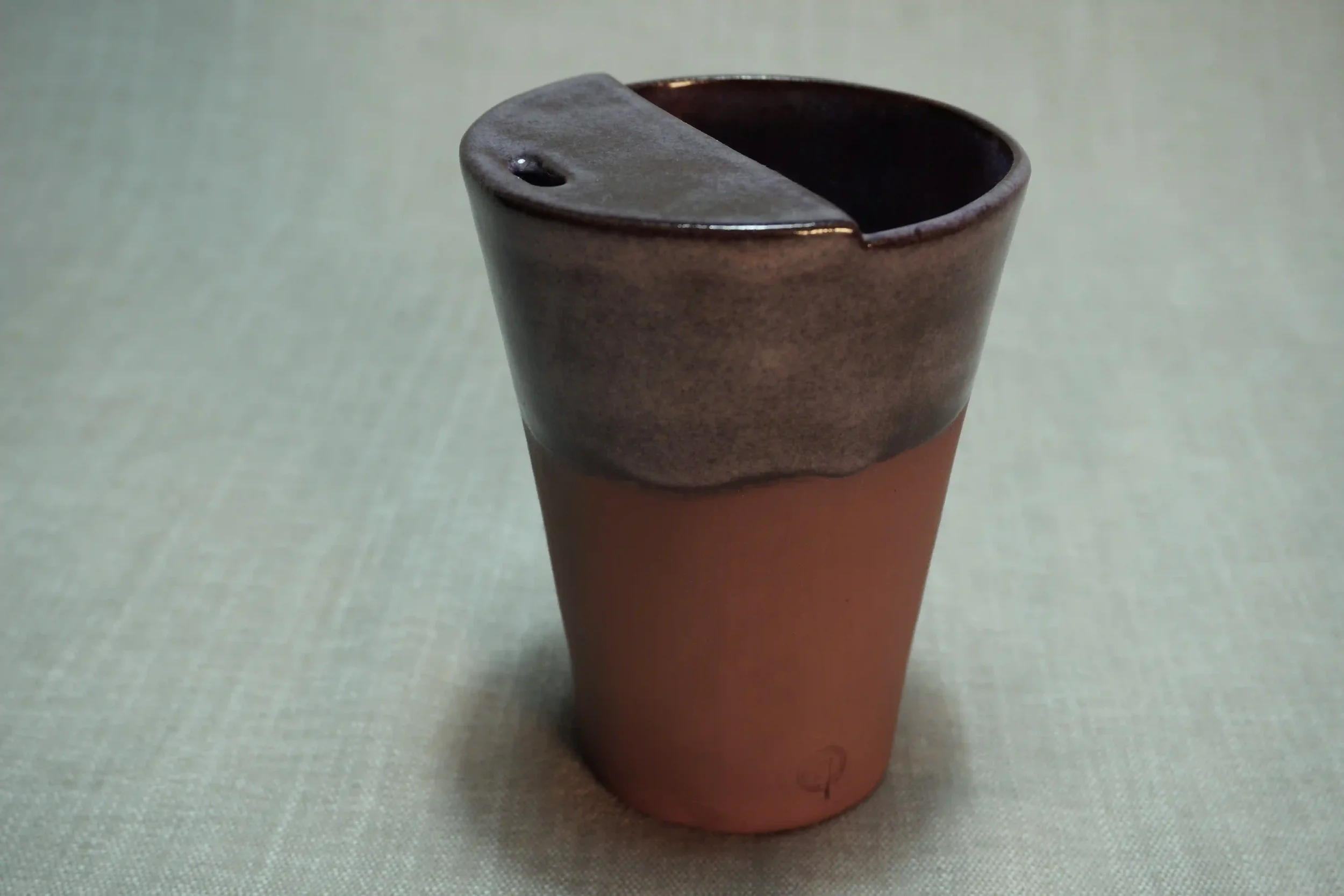 A brown ceramic cup with a rounded rim and a small hole, sitting on a light-colored surface.