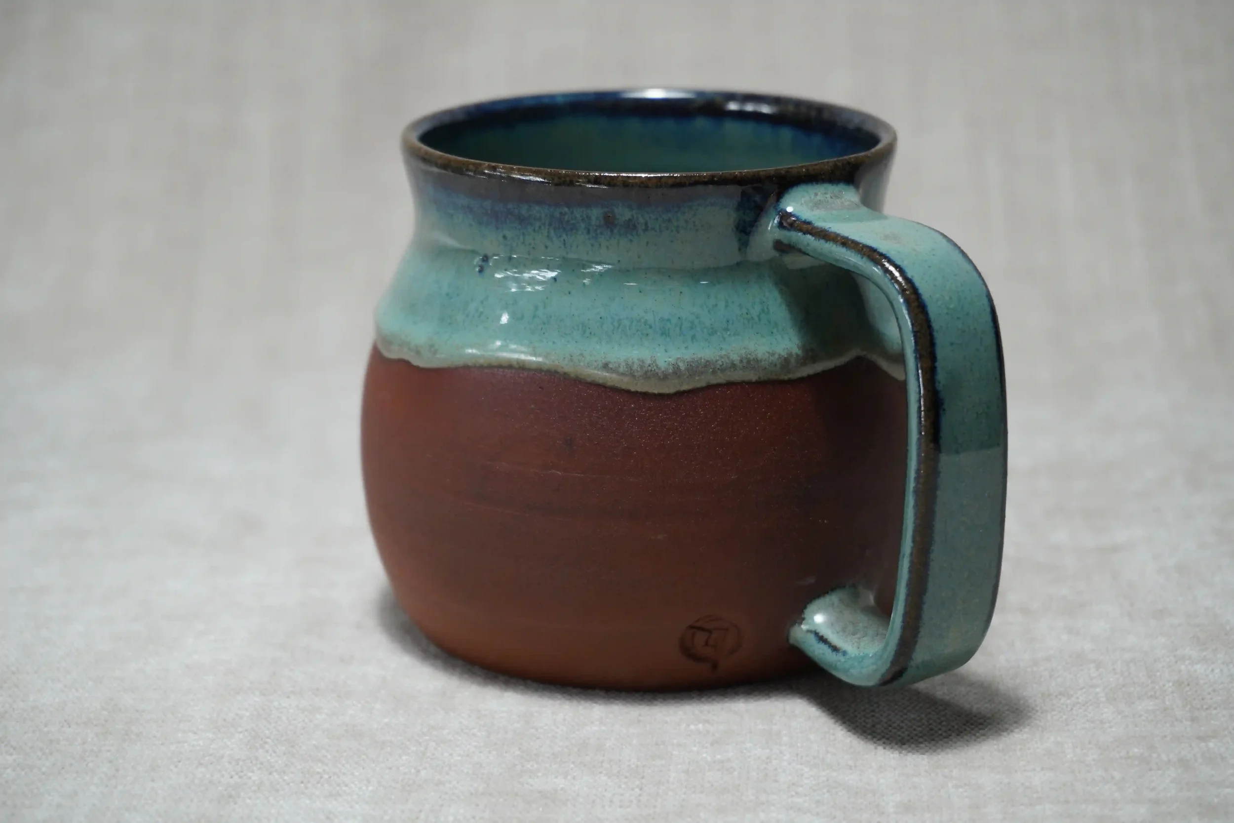 Ceramic belly mug with a matte terracotta brown bottom, glazed blue-green top, and a handle, placed on a light-colored surface.