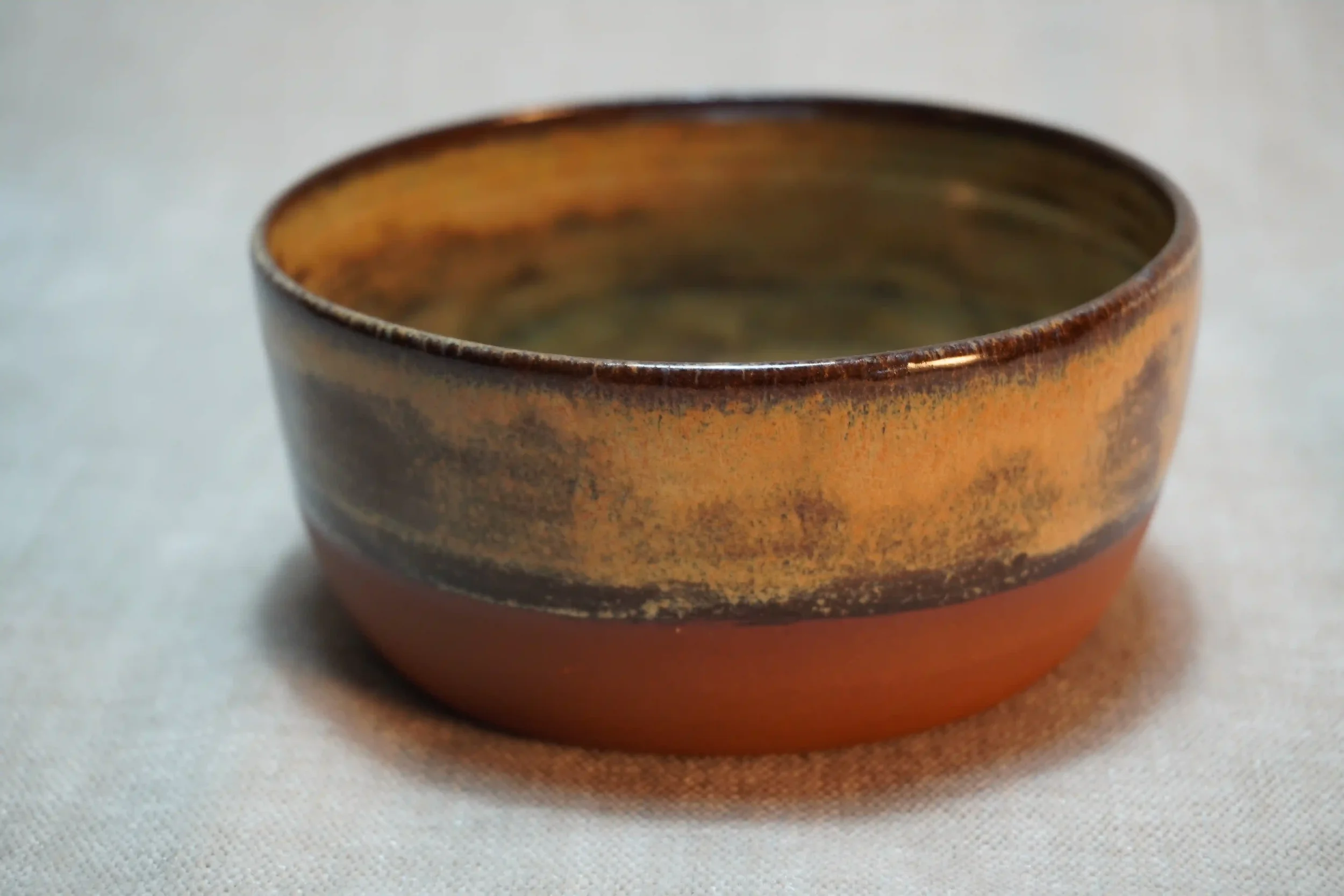 A small, round, handcrafted ceramic bowl with a honey-brown glaze and terracotta, earthy appearance, placed on a neutral surface.
