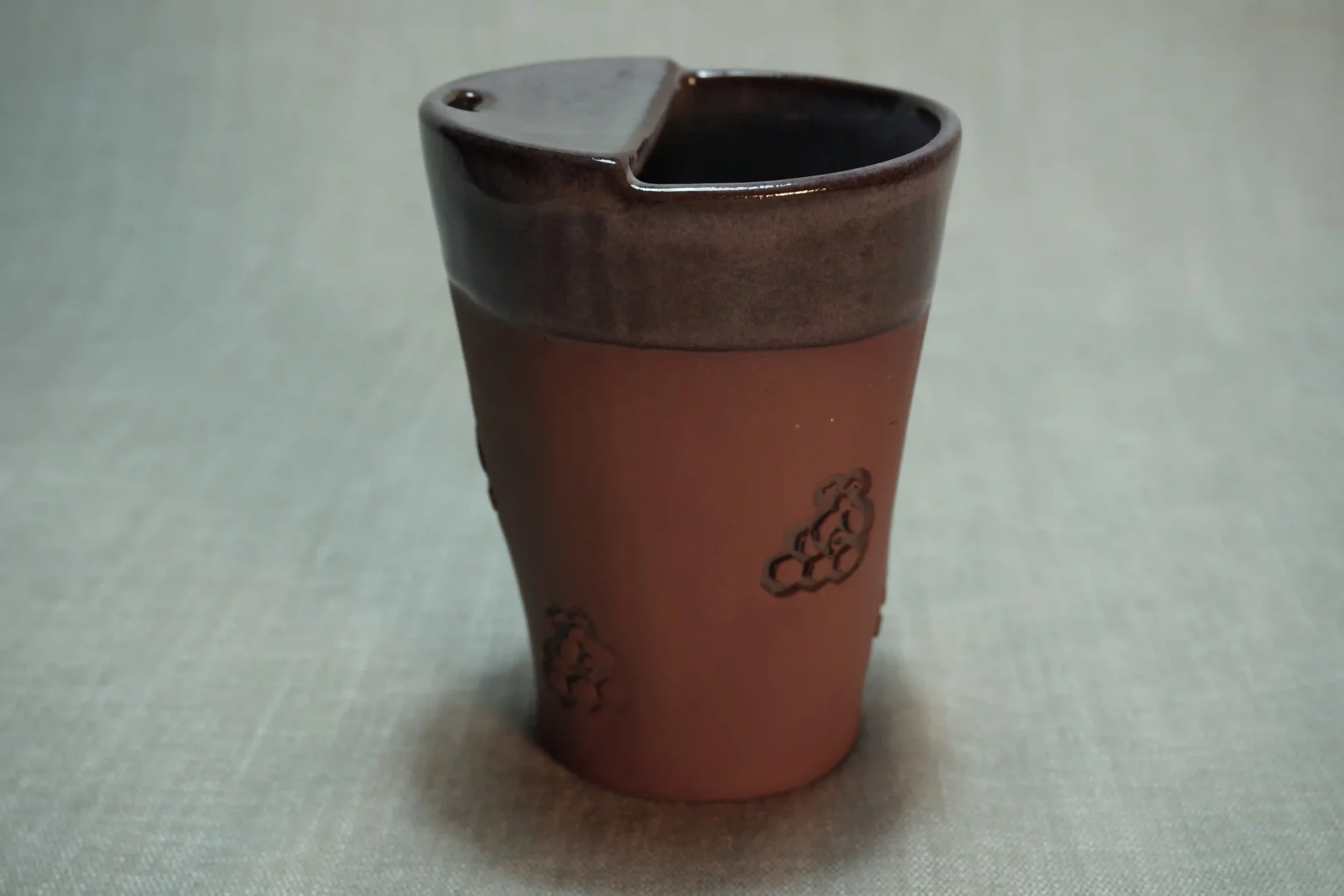A ceramic travel mug with a purple glaze, a terracotta brown base, and a decorative bee and honeycomb design on the outside, placed on a light-colored surface.