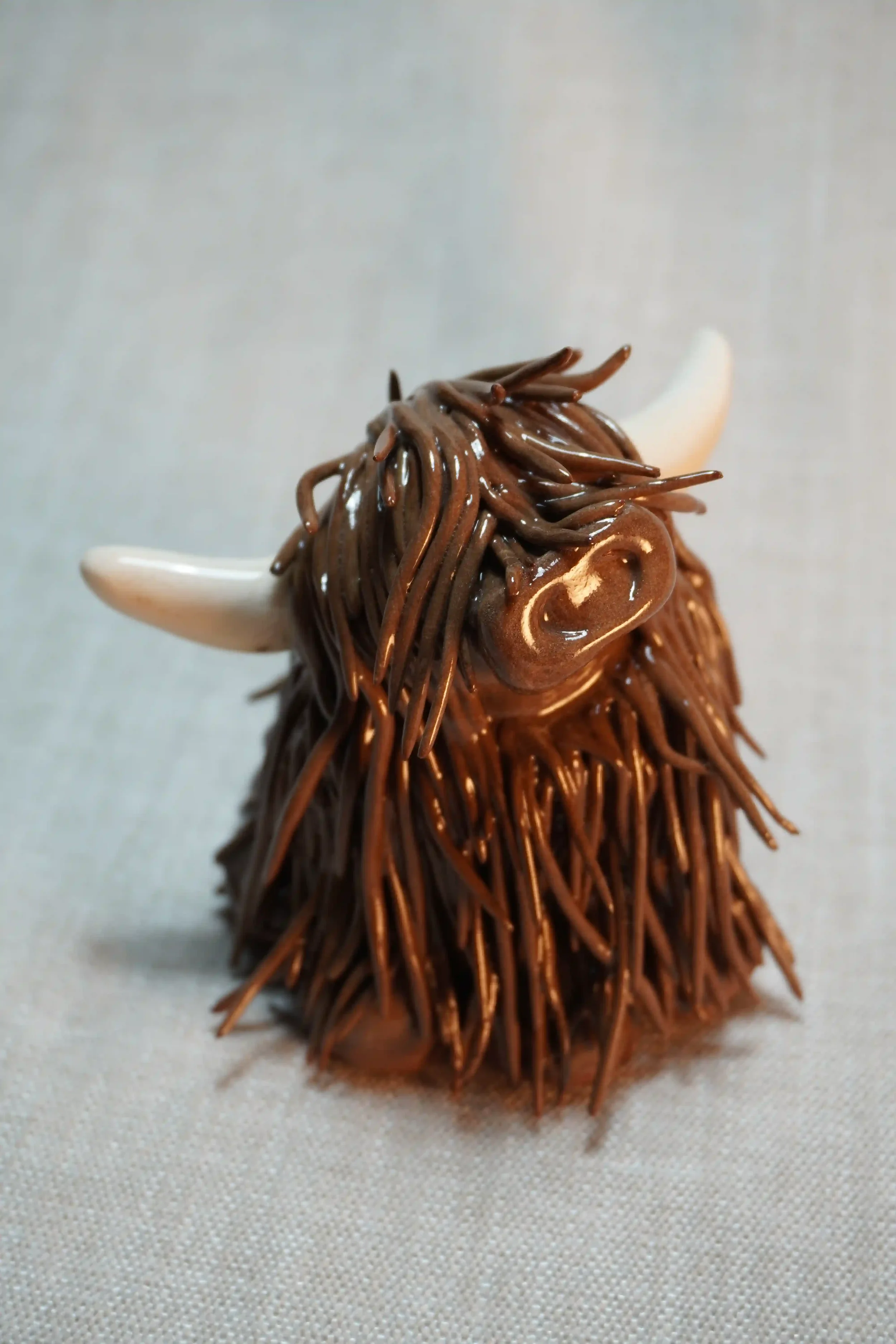 Front view of a handmade ceramic sculpture of a Highland Cow ornament with dark brown-coloured hair and beige horns.