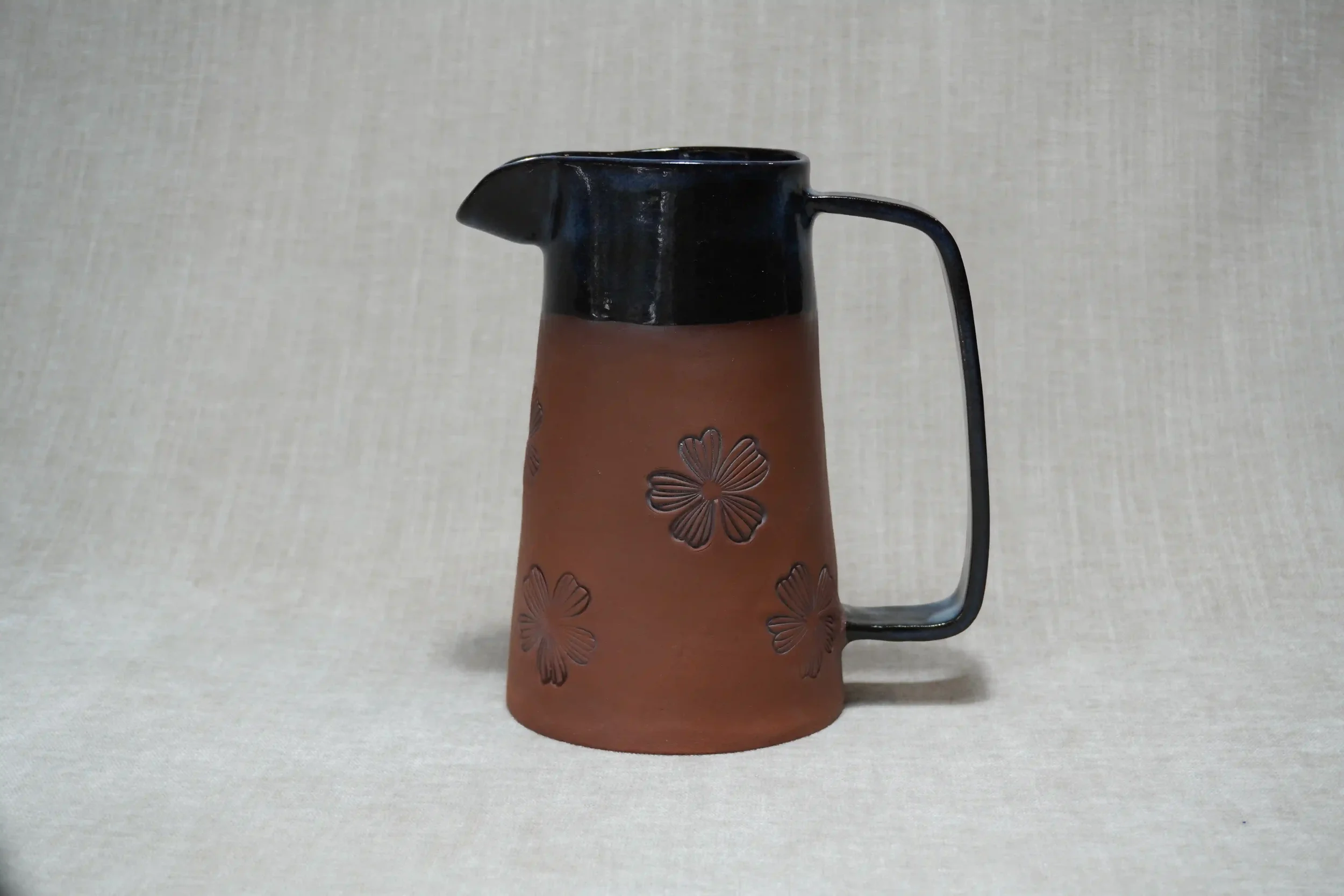 A ceramic pottery pitcher with a terracotta brown base and black glazed top, decorated with a floral pattern, and a black handle.