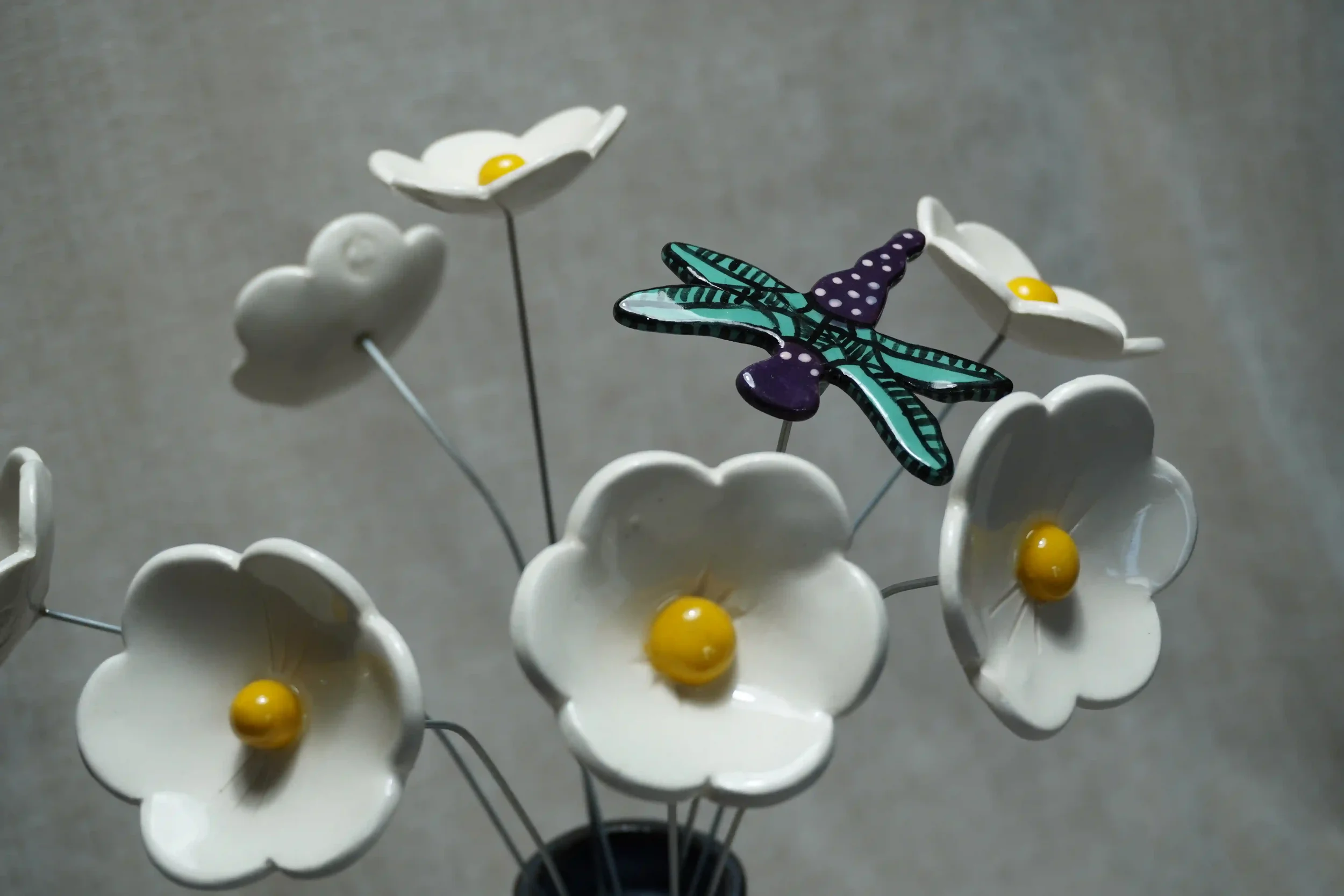 A close-up of a colorful ceramic dragonfly with teal and black wings and a purple body with white speckles, set amongst white ceramic daisies with yellow centres. All mounted on thin metal stems, against a plain gray background
