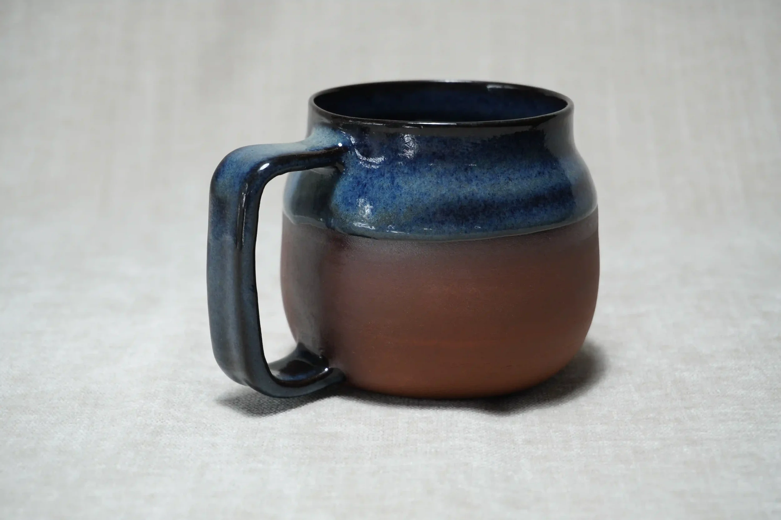 A ceramic mug with a blue glazed top rim and handle, and a matte terracotta brown lower body, placed on a plain light-colored surface.