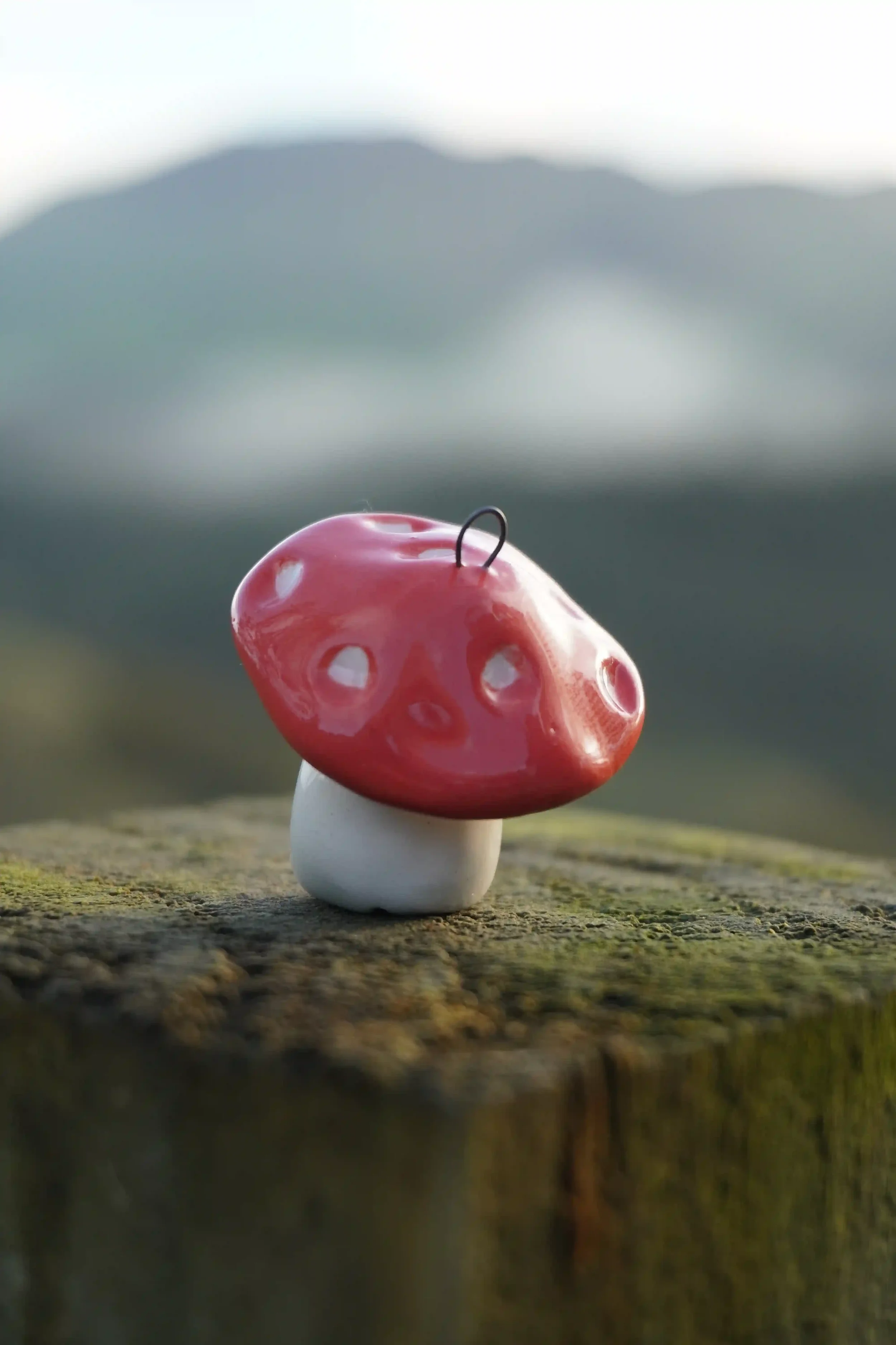A handmade pink and white ceramic mushroom-shaped ornaments with a hook on top, placed on a wooden surface with a blurred mountain landscape background.