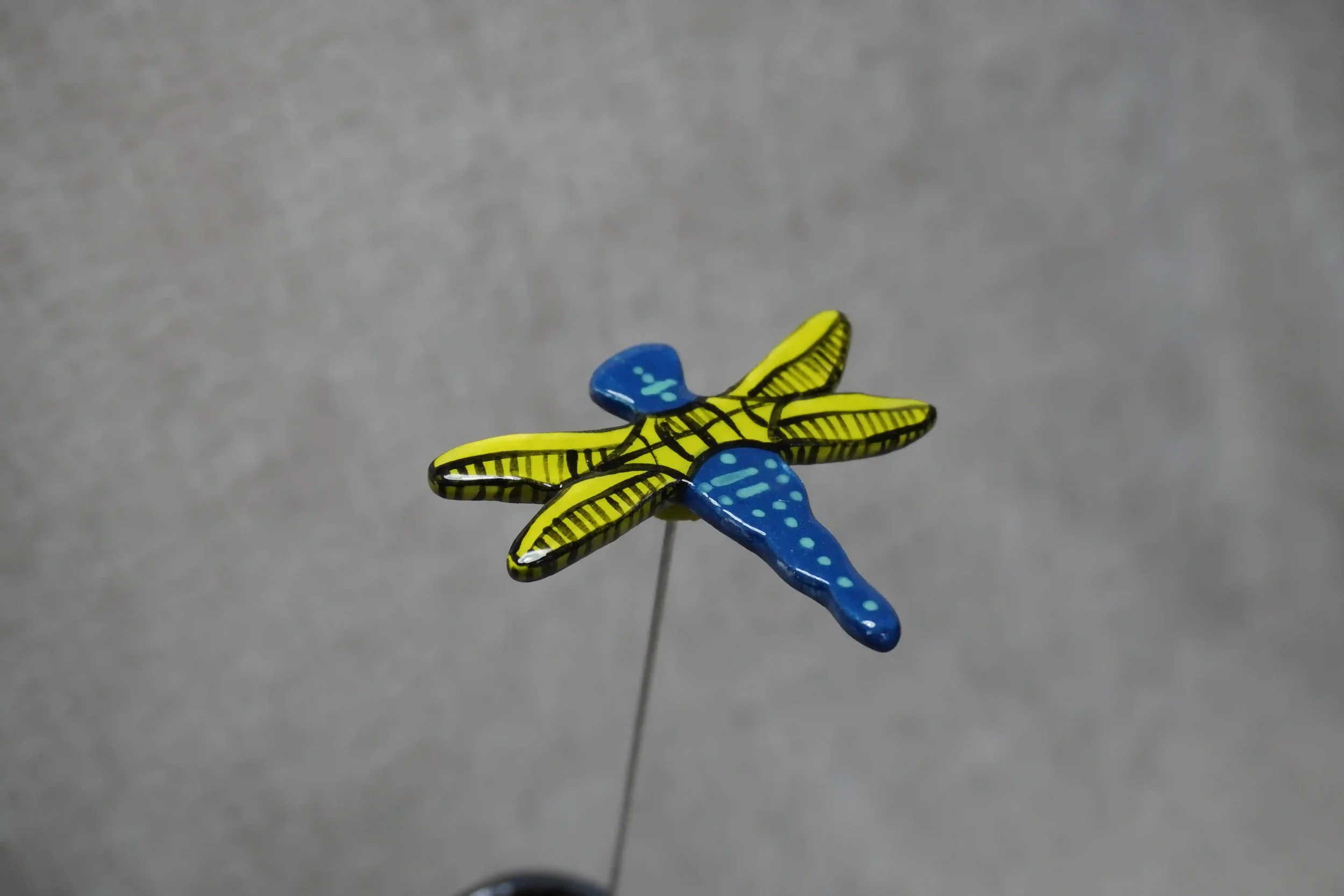 Colorful decorative dragonfly pin with yellow and blue wings and a blue body, mounted on a thin metal pin against a plain gray background.