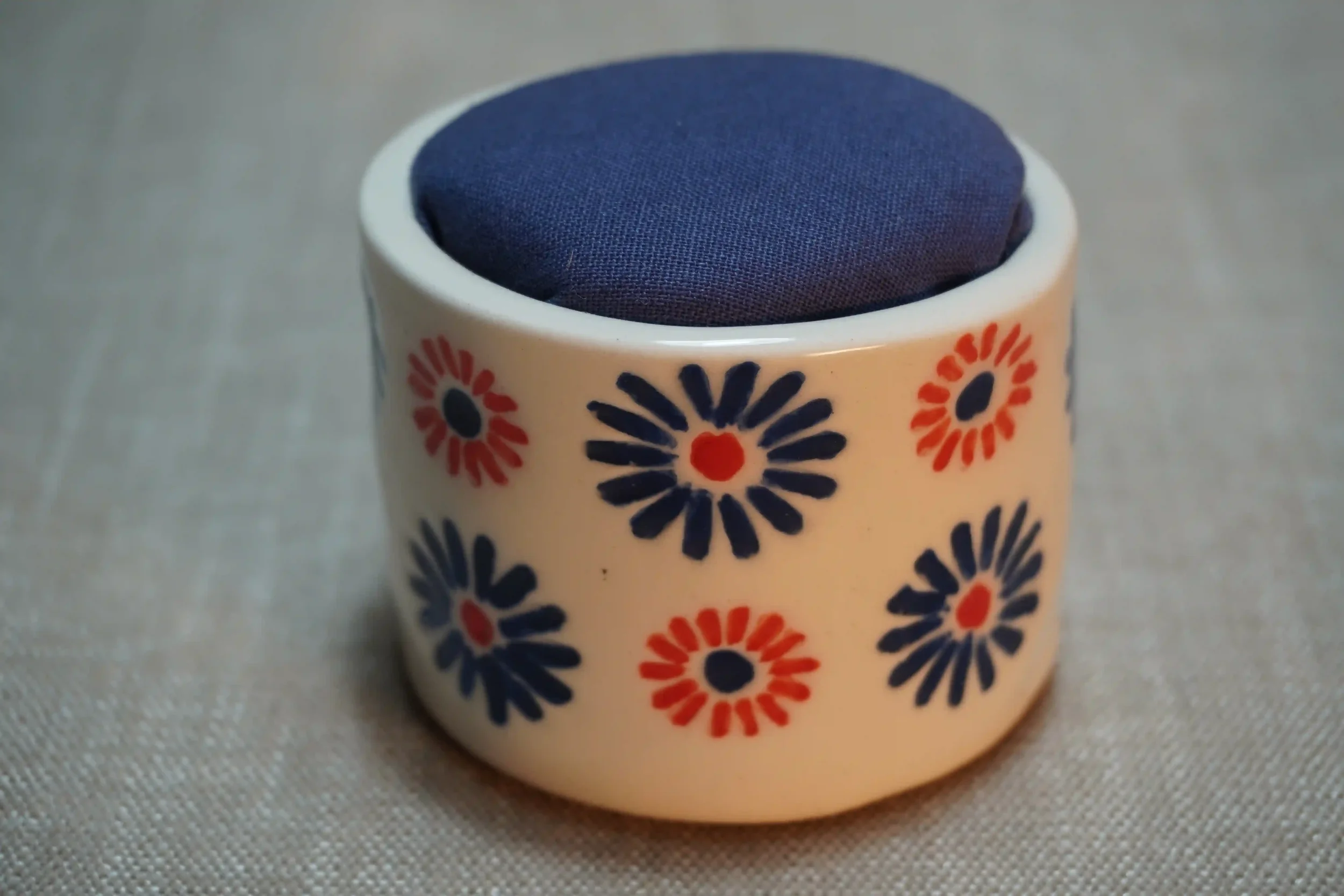 A ceramic pincushion with a red and blue firework pattern, filled with a blue cloth cushion. The pincushion is shown on a textured surface.