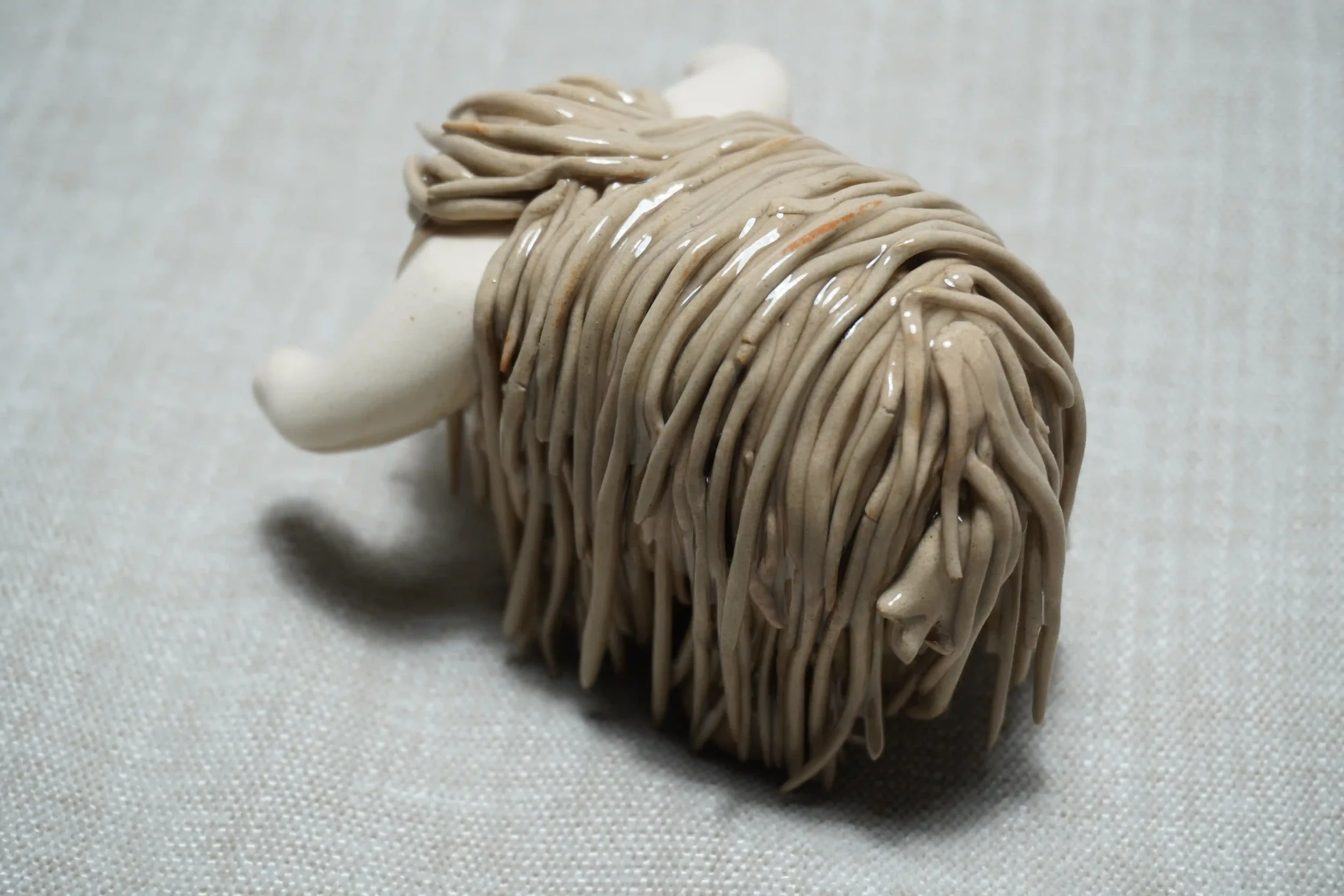 Quarter rear view of a cute handmade ceramic Highland Cow ornament with light brown coloured hair and white horns.