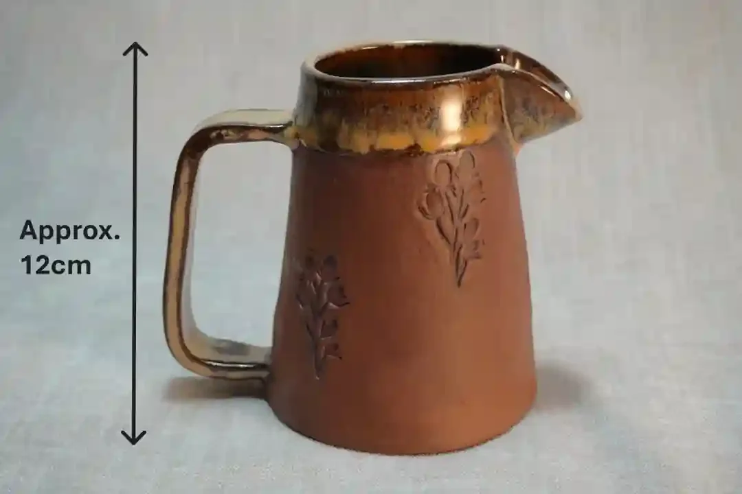Handmade ceramic pitcher (jug) with a handle, honey-coloured glaze, decorated with floral patterns, approximately 12 centimeters tall.
