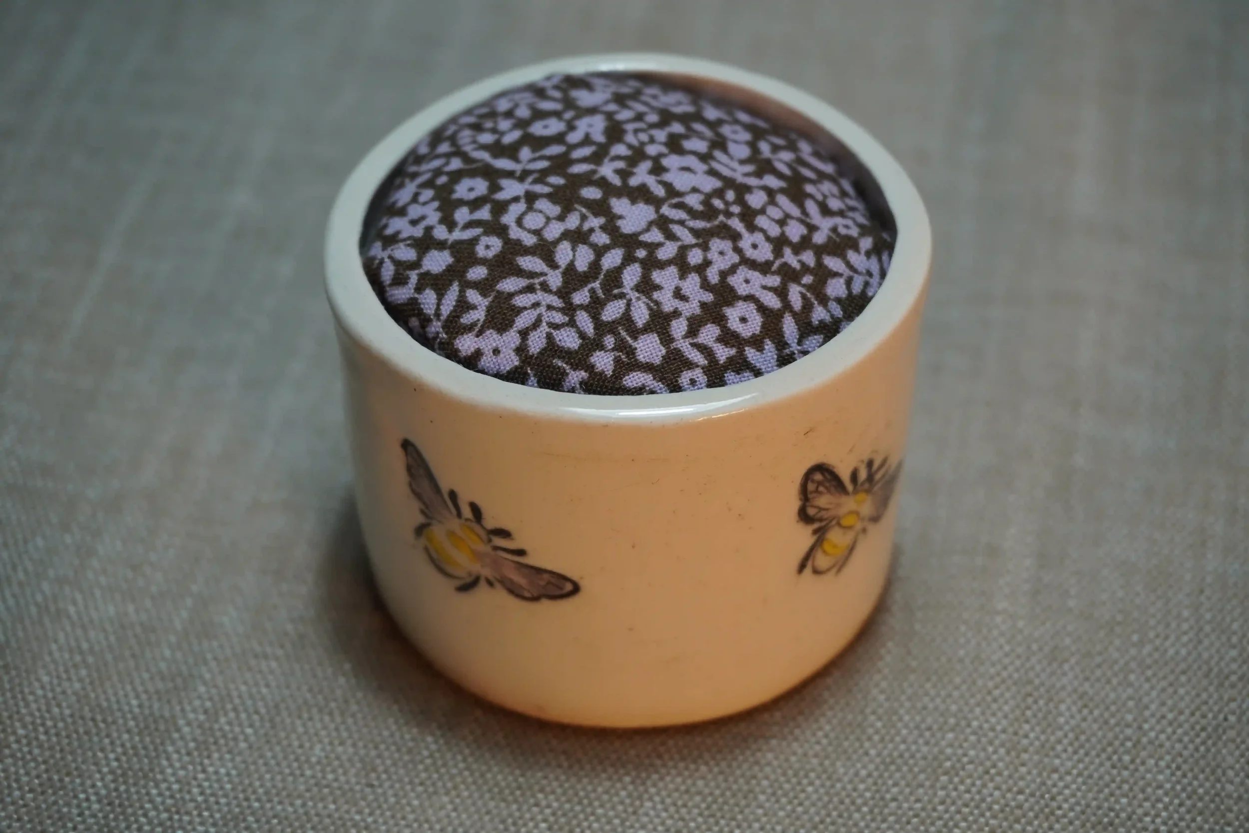 A ceramic pincushion with bee design, filled with a brown and pink floral patterned cushion. The pincushion is shown on a textured surface.