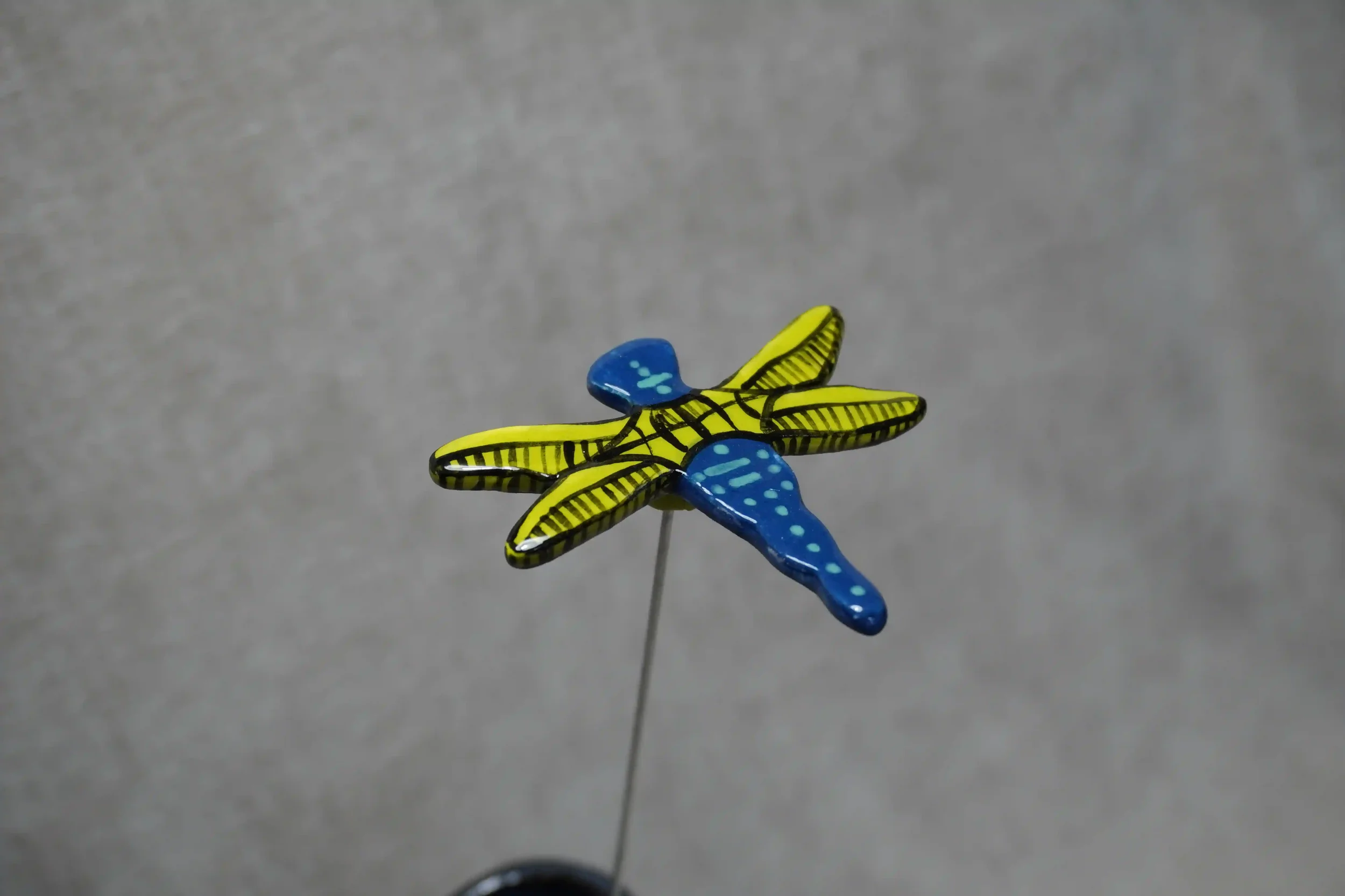Colorful ceramic dragonfly with yellow-green wings and a blue speckled body, mounted on a thin metal stem, against a plain gray background.