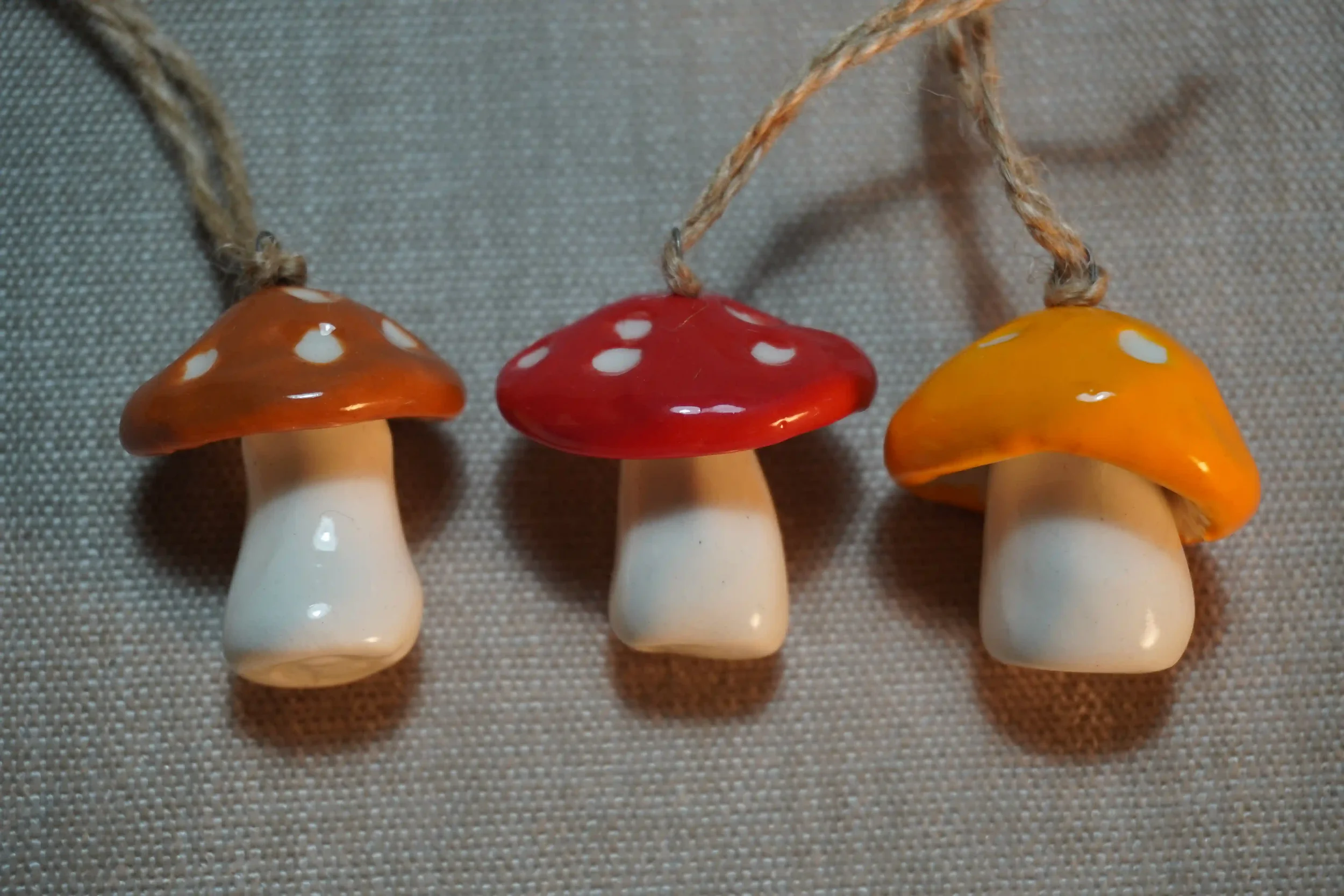 Three mushroom-shaped ceramic ornaments in brown, yellow and red with white spots, and with string loops hanging on a fabric background.