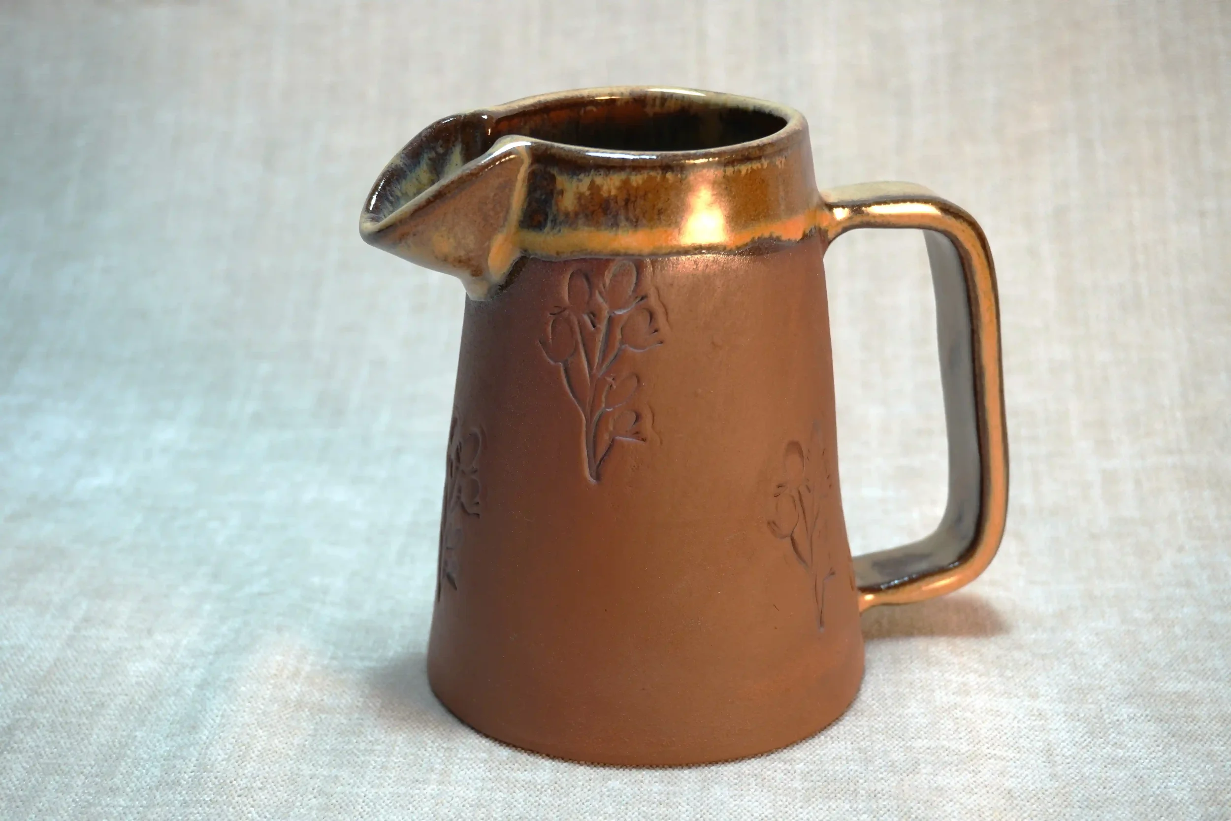 A ceramic pitcher with an earthy terracotta brown color and honey-coloured glaze, decorated with embossed floral designs, featuring a large handle and a spout.