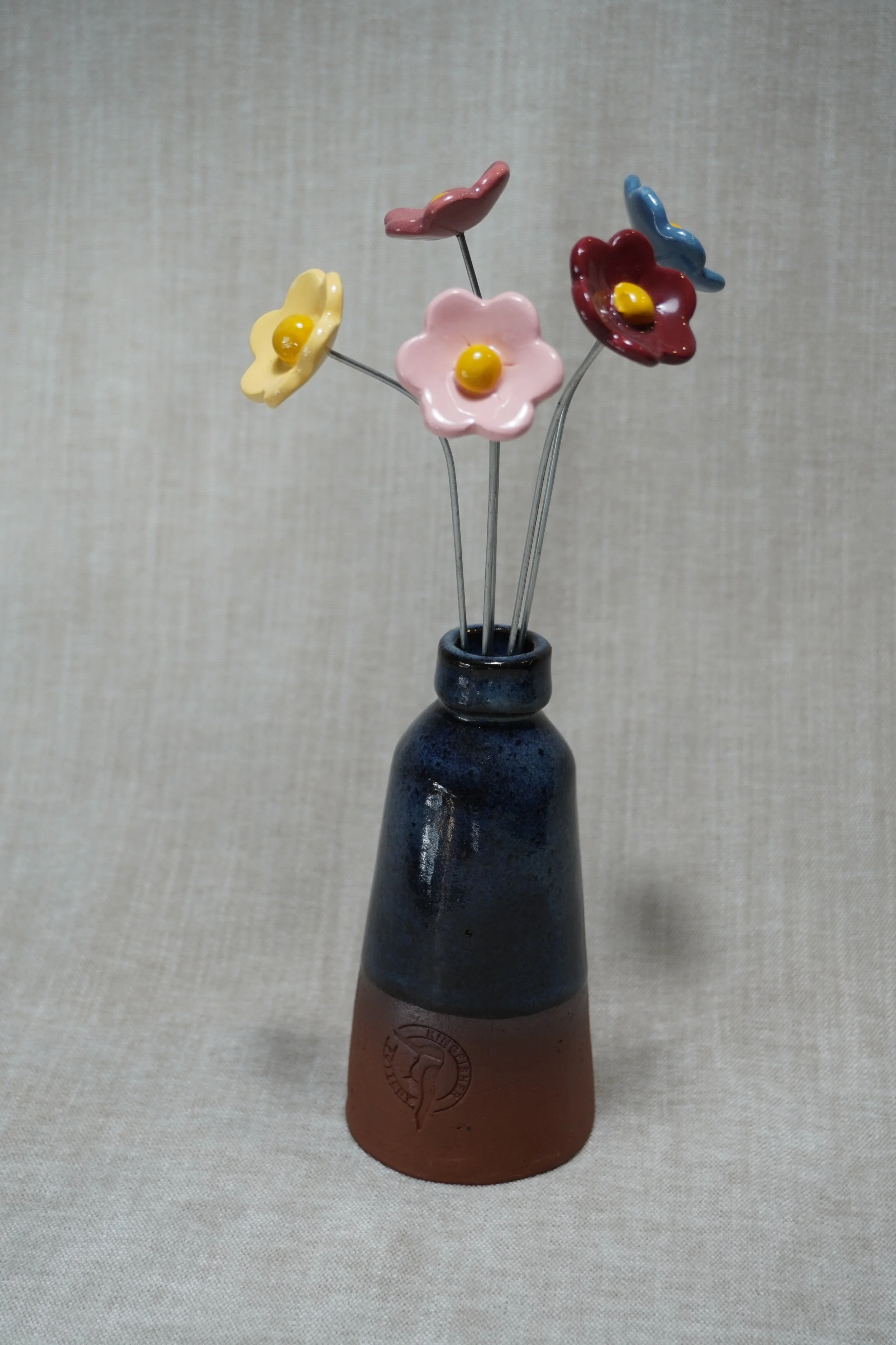 A ceramic vase with a wide, rounded terracotta base, narrow neck, and a blue glaze, holding five colorful ceramic flowers in light pink, medium pink and dark pink, blue and yellow with yellow centers set against a beige textured background.