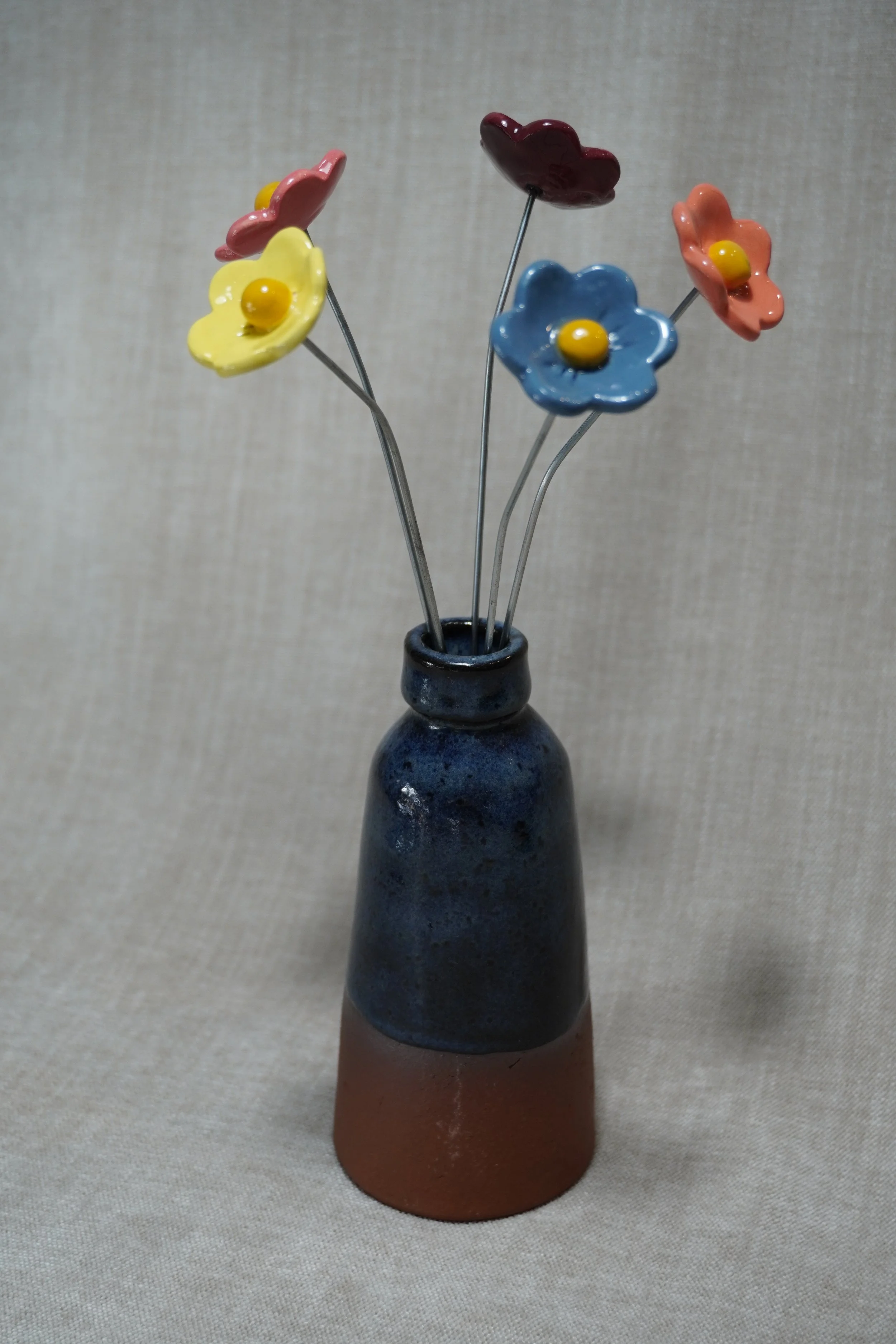 A ceramic vase with a wide, rounded terracotta base, narrow neck, and a blue glaze, holding five ceramic flowers in yellow, blue, orange, pink and maroon, with yellow centres and metal stems, displayed on a light neutral background.