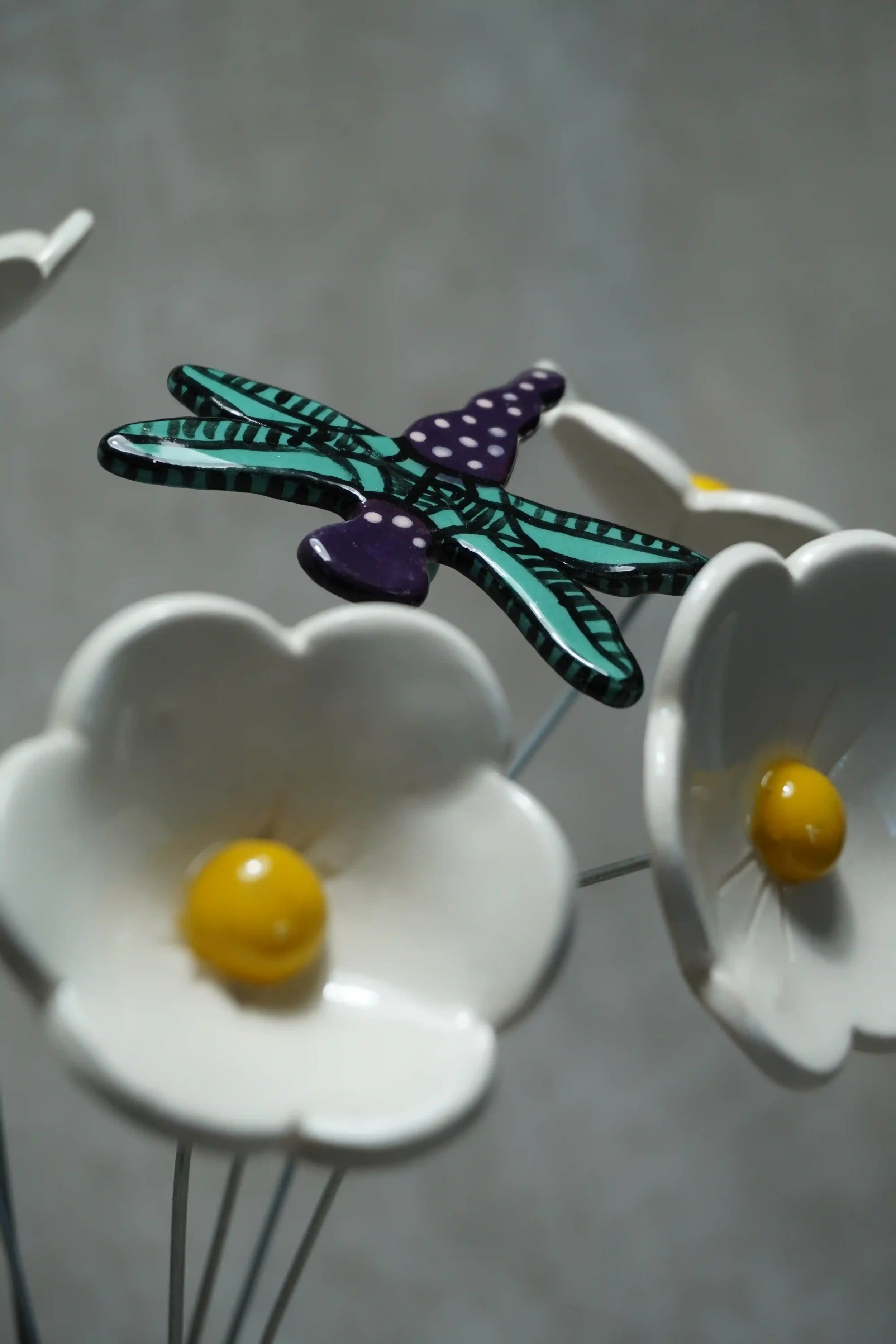 A close-up of a colorful ceramic dragonfly with teal and black wings and a purple body with white speckles, set amongst white ceramic daisies with yellow centres. All mounted on thin metal stems, against a plain gray background