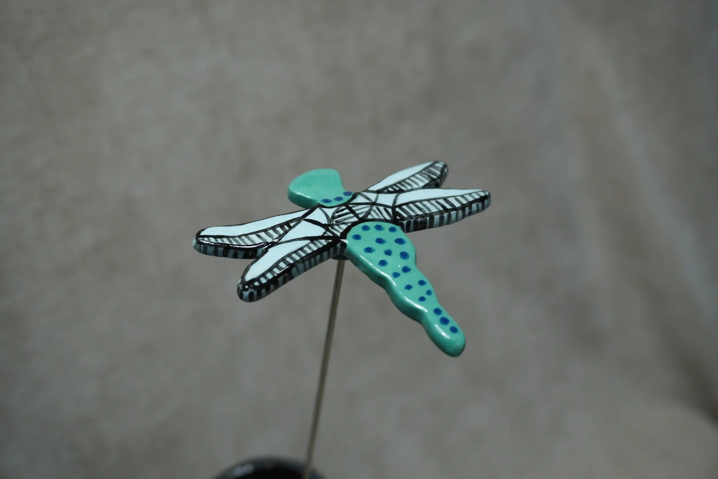 A small handmade ceramic dragonfly figurine with a turquoise body with blue spots, black and light blue wings , mounted on a metal stem against a plain gray background.