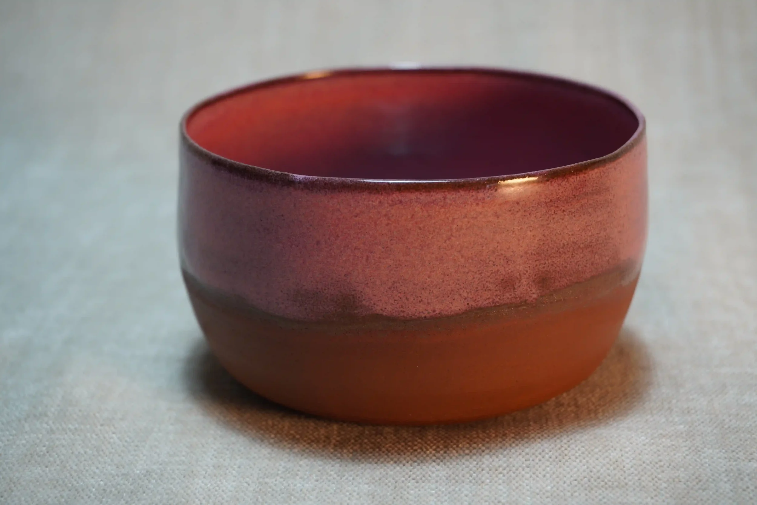 A small, round, handcrafted ceramic bowl with a pink glaze and terracotta brown base on a light-colored surface.